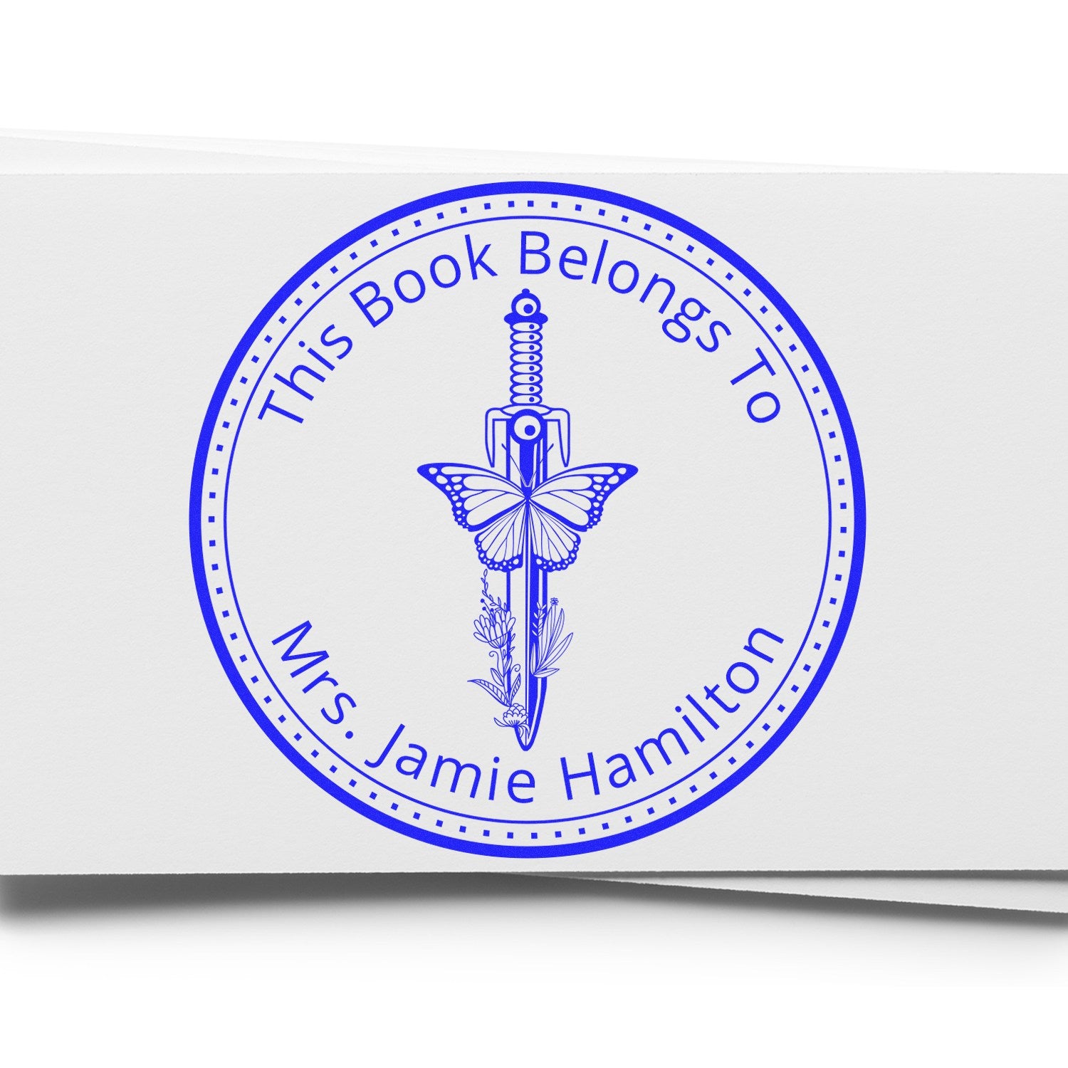 Blue circular book ownership stamp seal with sword and butterfly, reading This Book Belongs To Mrs. Jamie Hamilton, on white paper.