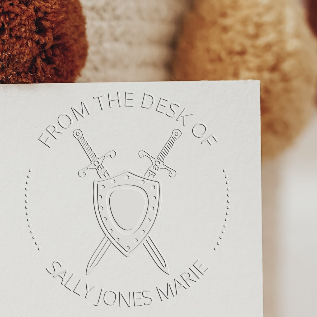 Custom white embossed desk seal stamp with crossed swords and shield on a warm brown background
