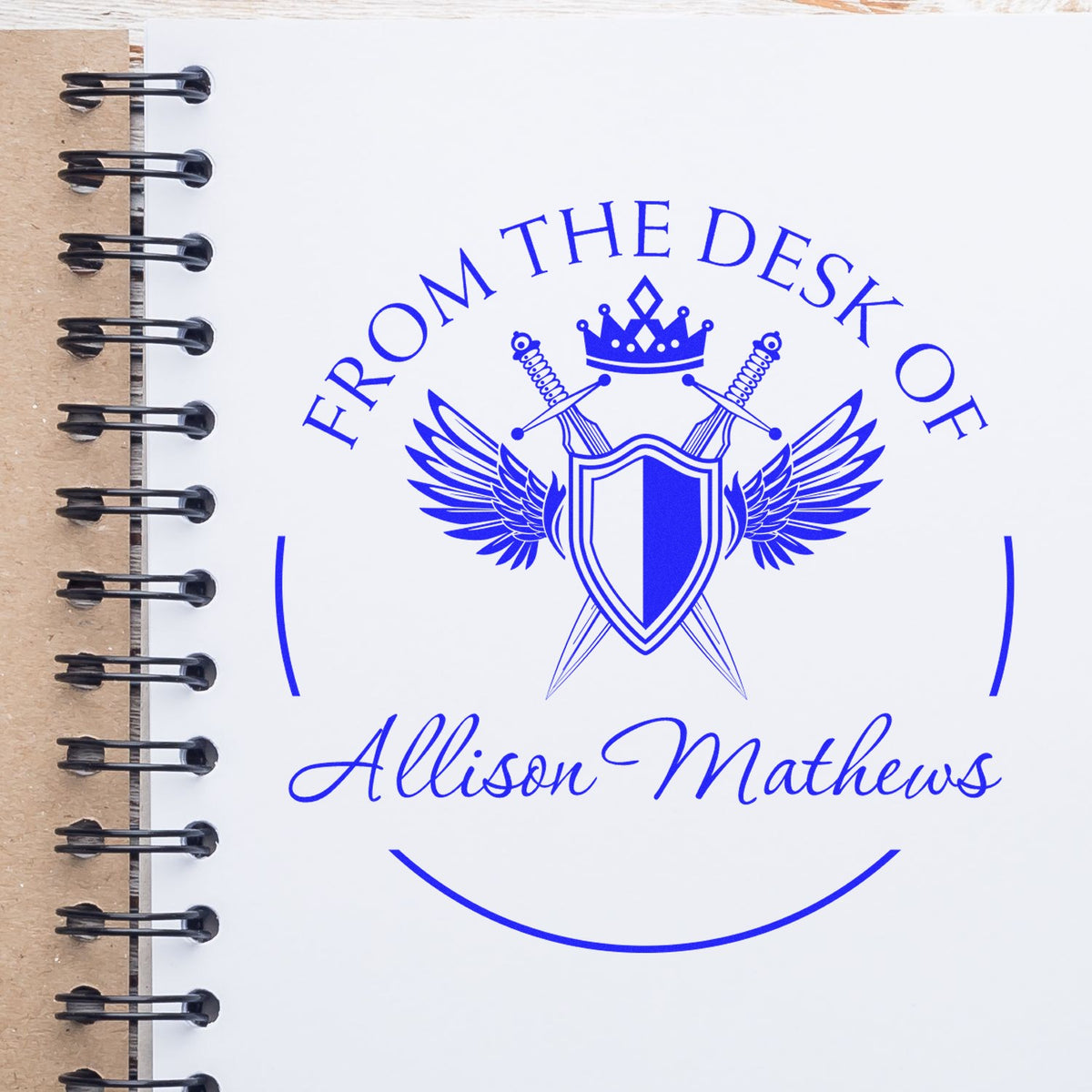 Blue stamp and seal on white spiral notebook, with crown, shield, wings, swords, and From the Desk of Allison Mathews.