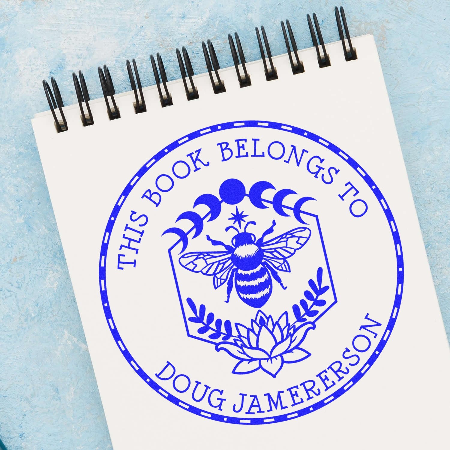 Blue custom bookplate stamp seal on a white spiral notebook, featuring a bee, moon phases, lotus, and This Book Belongs to Doug Jamerson.