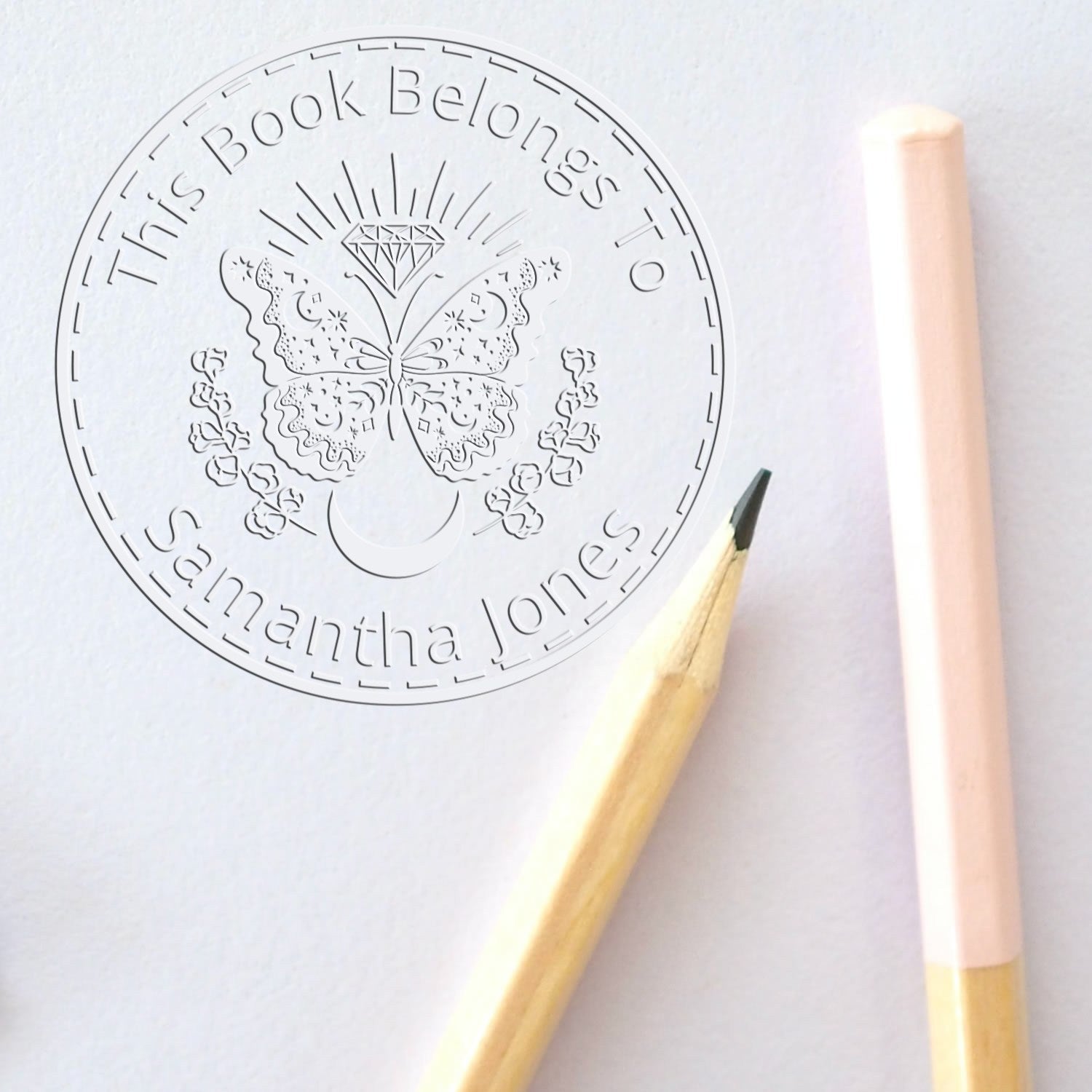 White embossed stamp seal with butterfly and diamond, beside pale pink and wooden pencils on white paper.