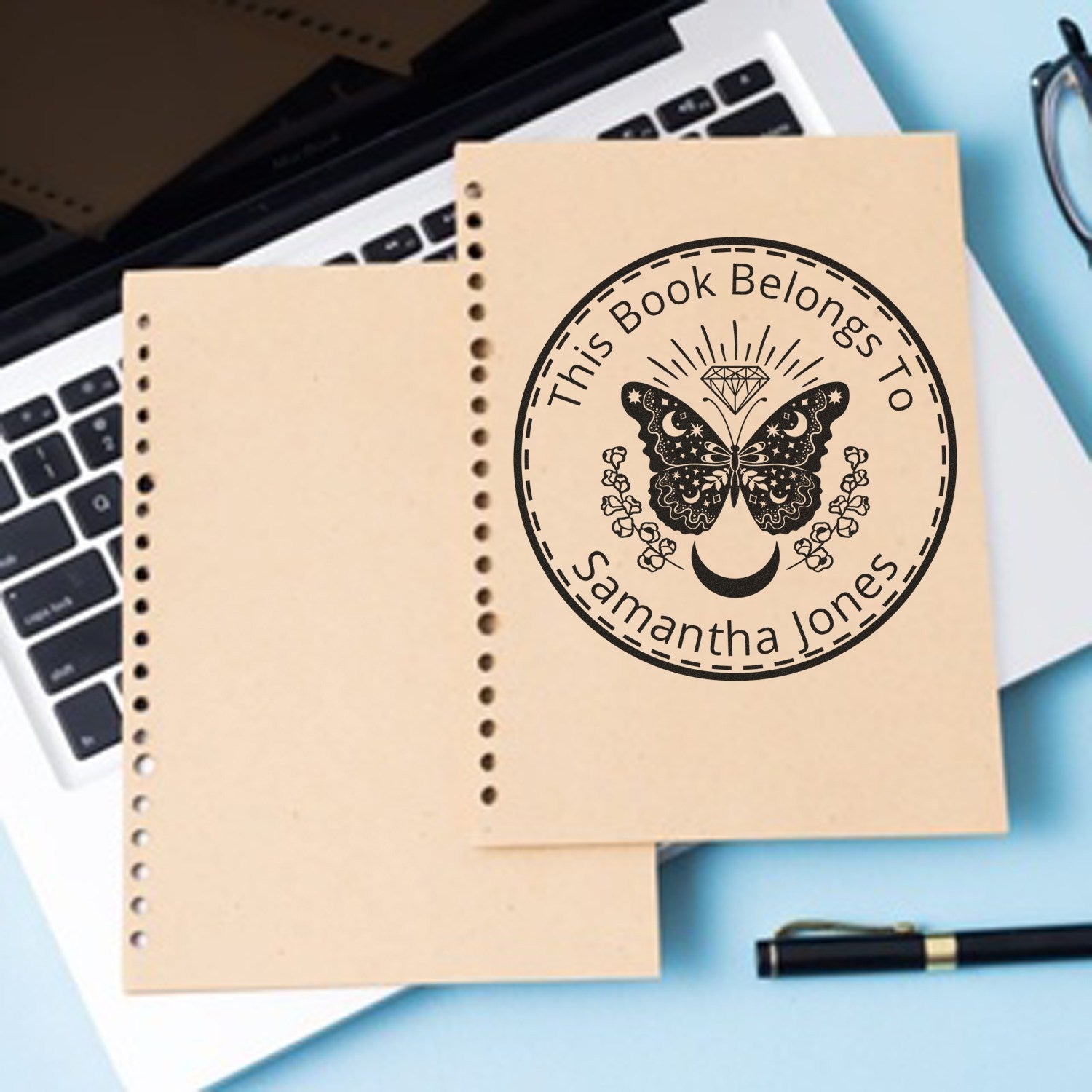 Personalized black book stamp and seal on tan paper, featuring butterfly, moon, diamond, and floral design on a laptop desk.
