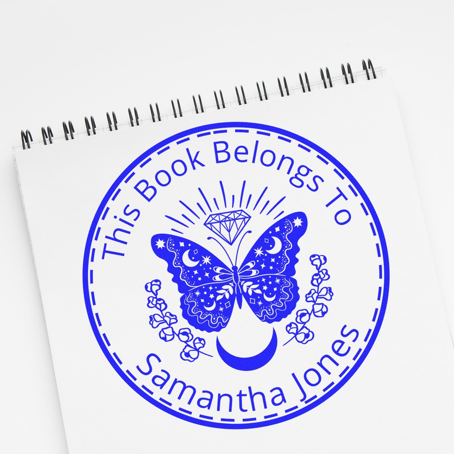 Blue personalized book stamp seal on notepad with butterfly, diamond, moon and flowers, labeled This Book Belongs To Samantha Jones.