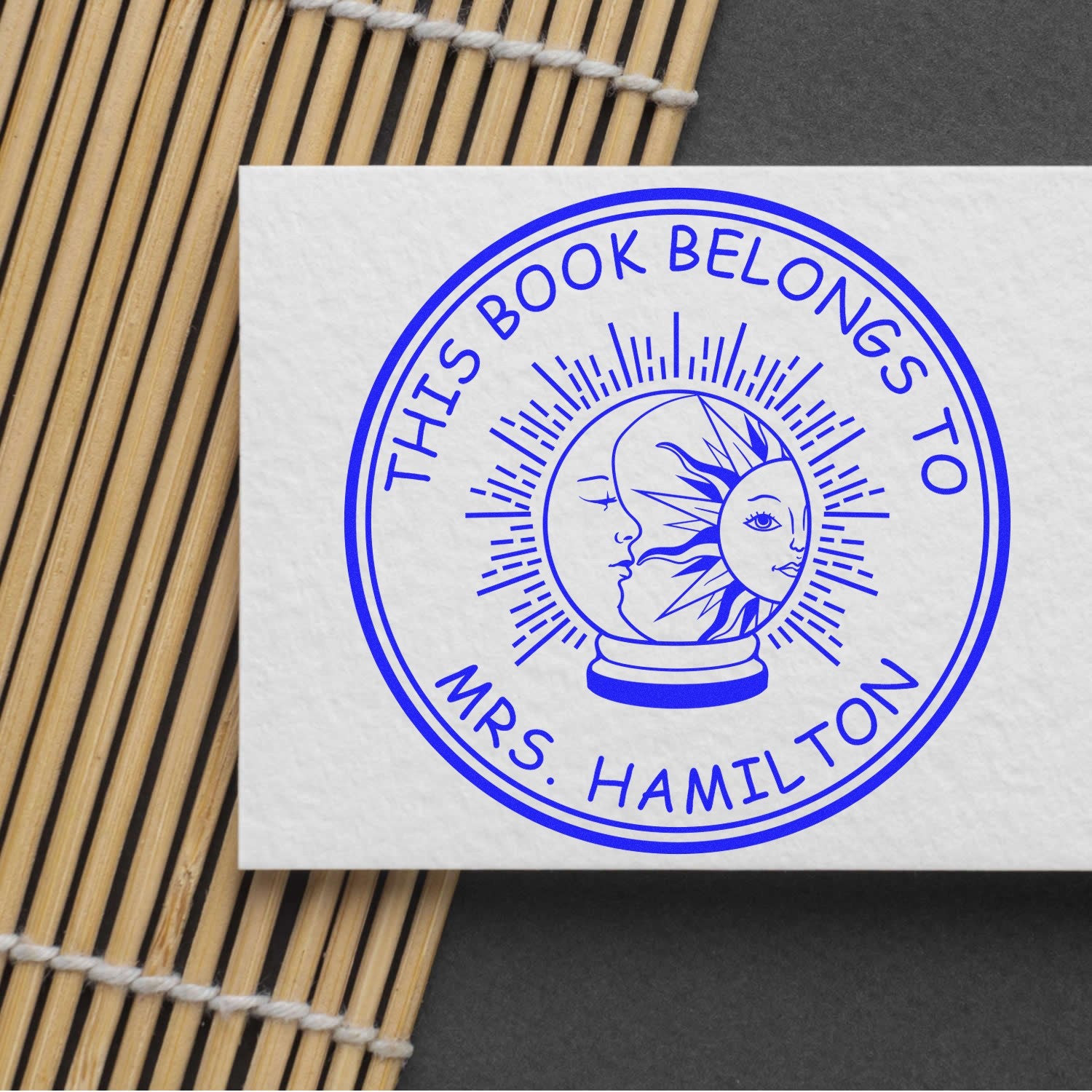 Blue round self-inking stamp seal reading This Book Belongs to Mrs. Hamilton with moon-and-sun design on white paper.