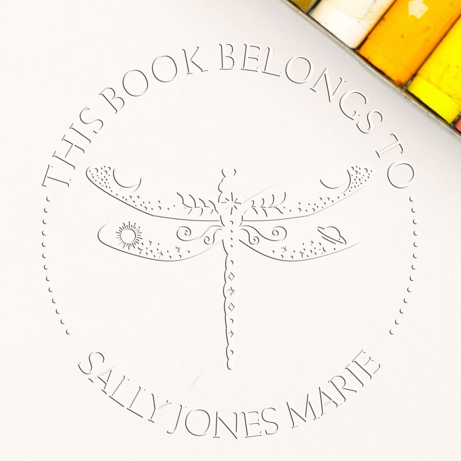 Embossed book stamp seal with dragonfly, black lettering on white, yellow accents, reading “This Book Belongs to Sally Jones Marie.”