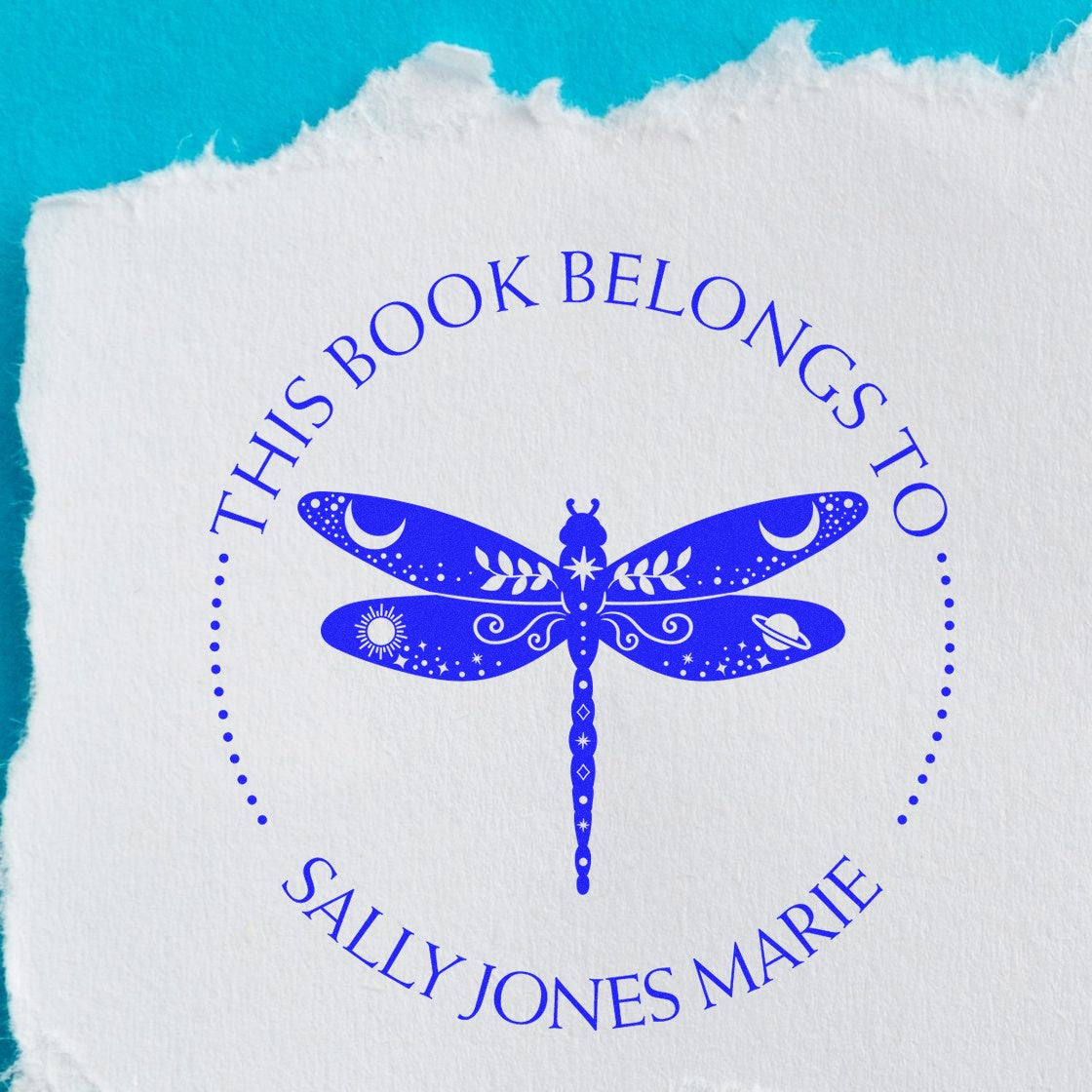 Blue custom book stamp and seal with dragonfly, moon and stars, reading This Book Belongs to Sally Jones Marie on white paper.