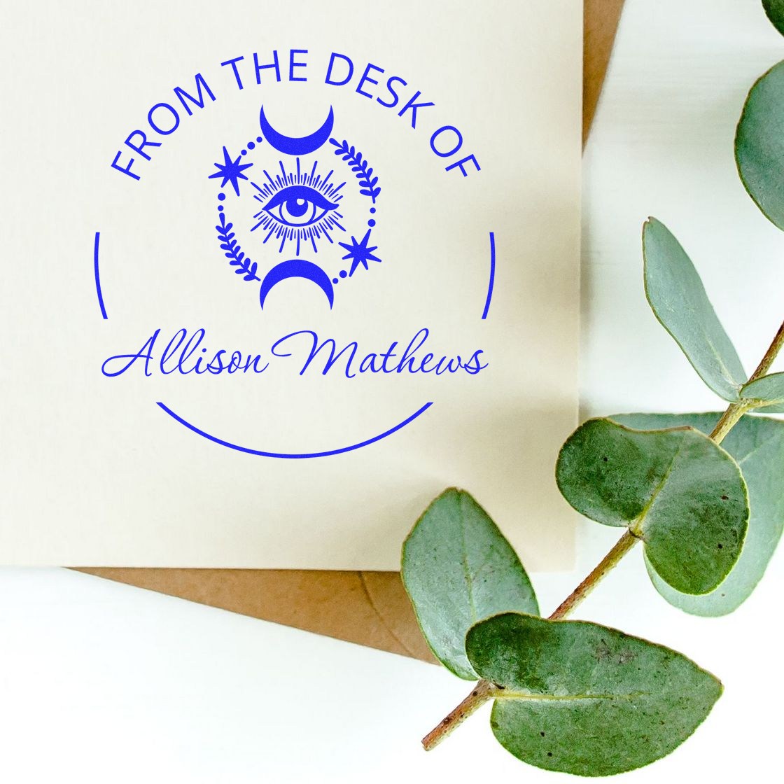 Blue custom desk stamp and seal for Allison Mathews on cream paper, featuring a moon-and-eye emblem and eucalyptus leaves.