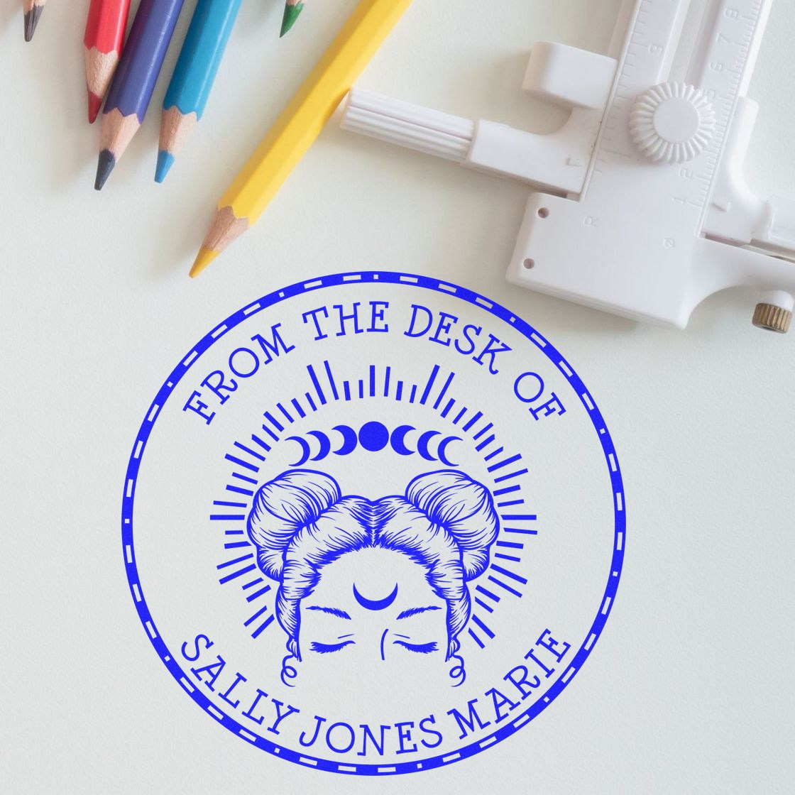 Blue circular custom stamp seal with moon goddess design, beside colored pencils and a white embossing tool on a white desk.