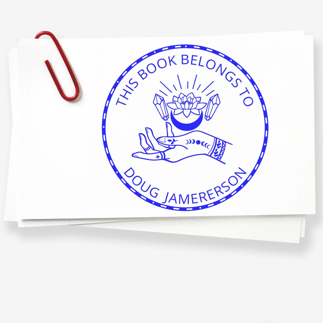 Blue custom book ownership stamp seal on white paper, with lotus, hand design, and red paperclip.