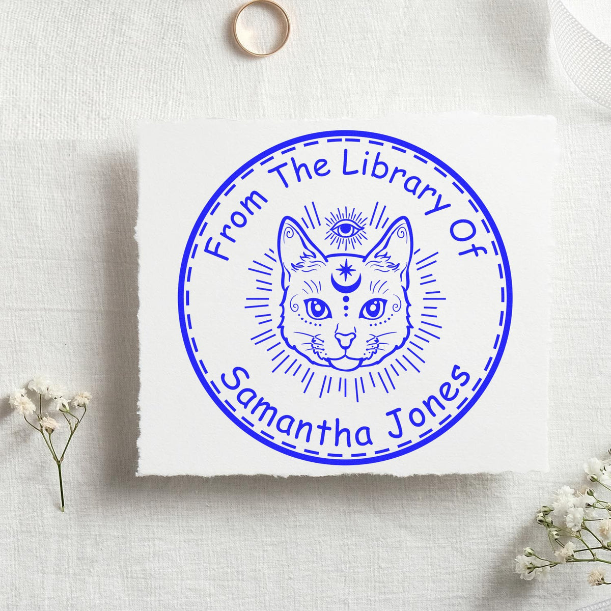 Blue circular library stamp seal with cat face, reading From The Library Of Samantha Jones, on white fabric.