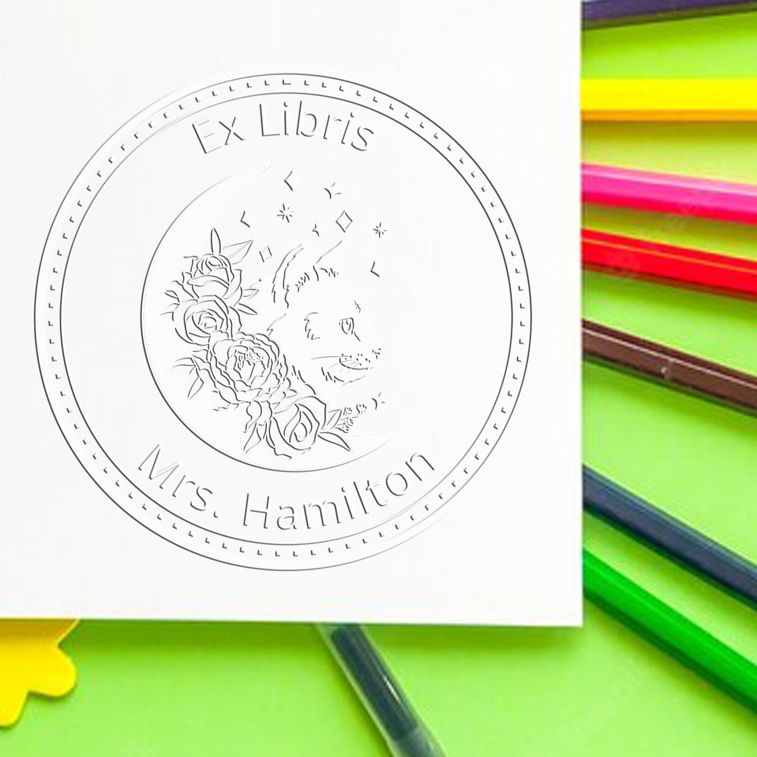 Circular Ex Libris stamp seal with floral wolf design and Mrs. Hamilton text on white paper, beside bright rainbow pencils.