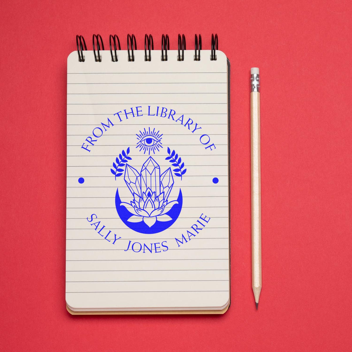 Blue custom library stamp and seal reading From the Library of Sally Jones Marie on a spiral notebook, with pencil on a red background.