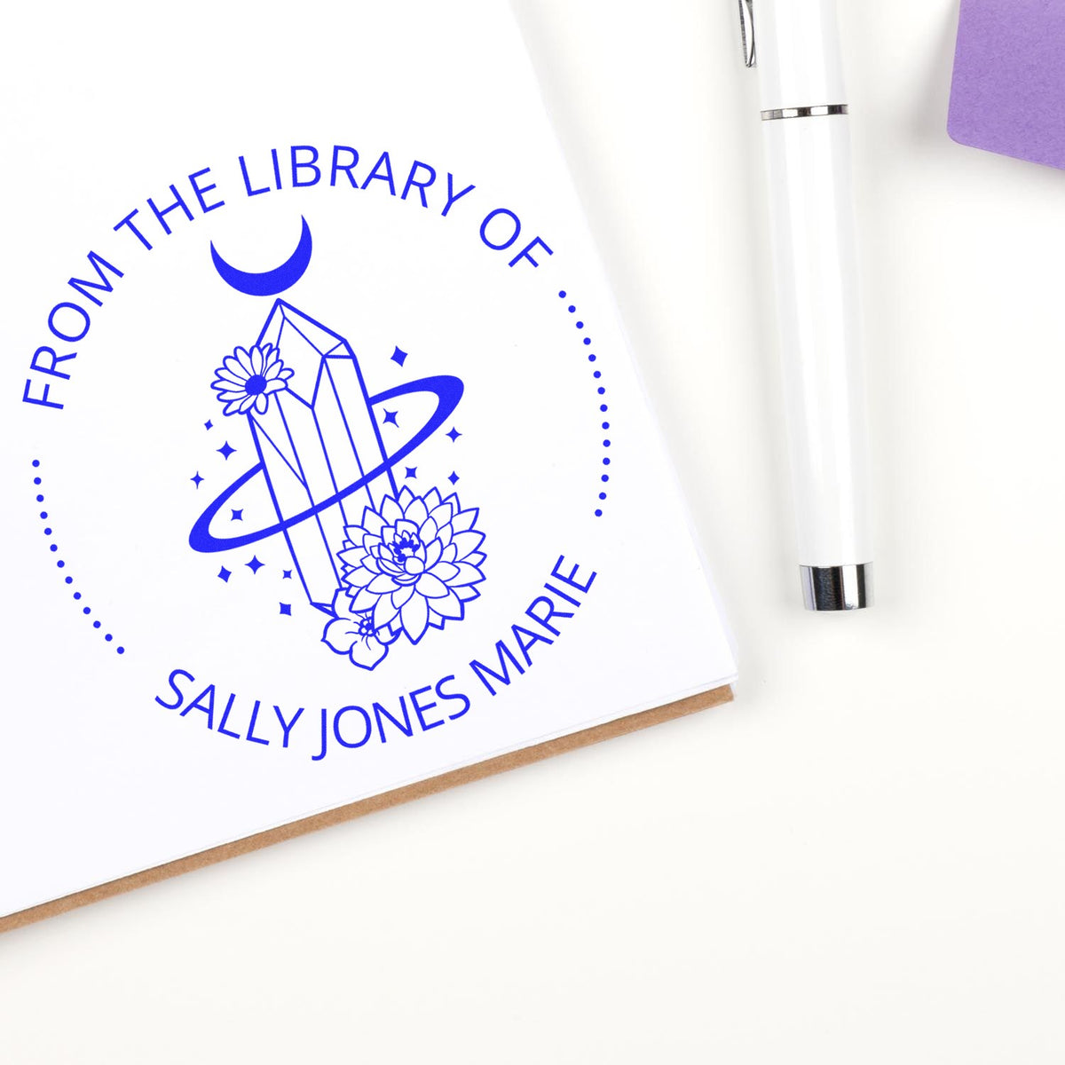 Blue custom library stamp seal with crystal and floral design on white paper, beside a white pen and purple accent.