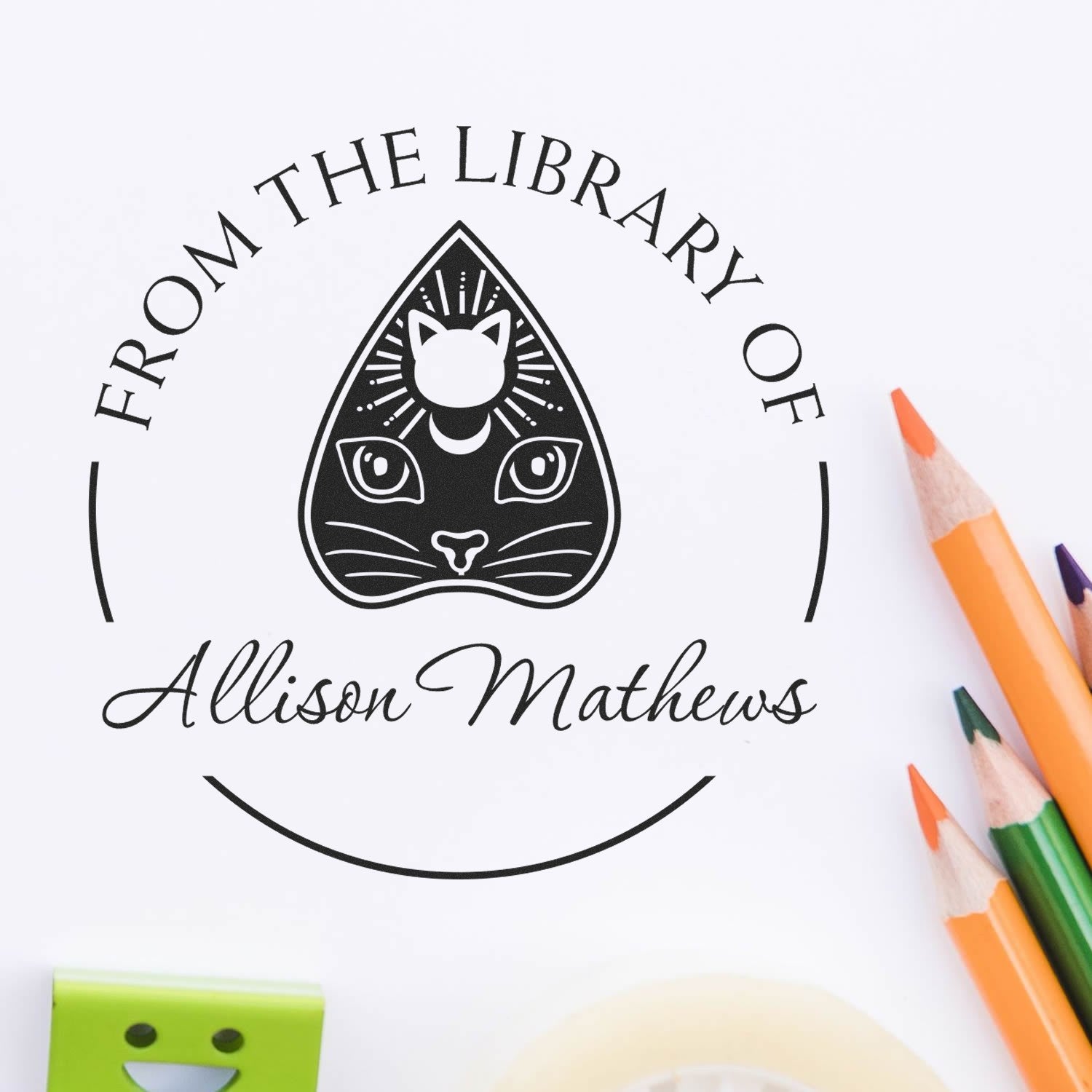 Custom black library stamp and seal with cat logo and Allison Mathews text, shown with colored pencils on white paper.