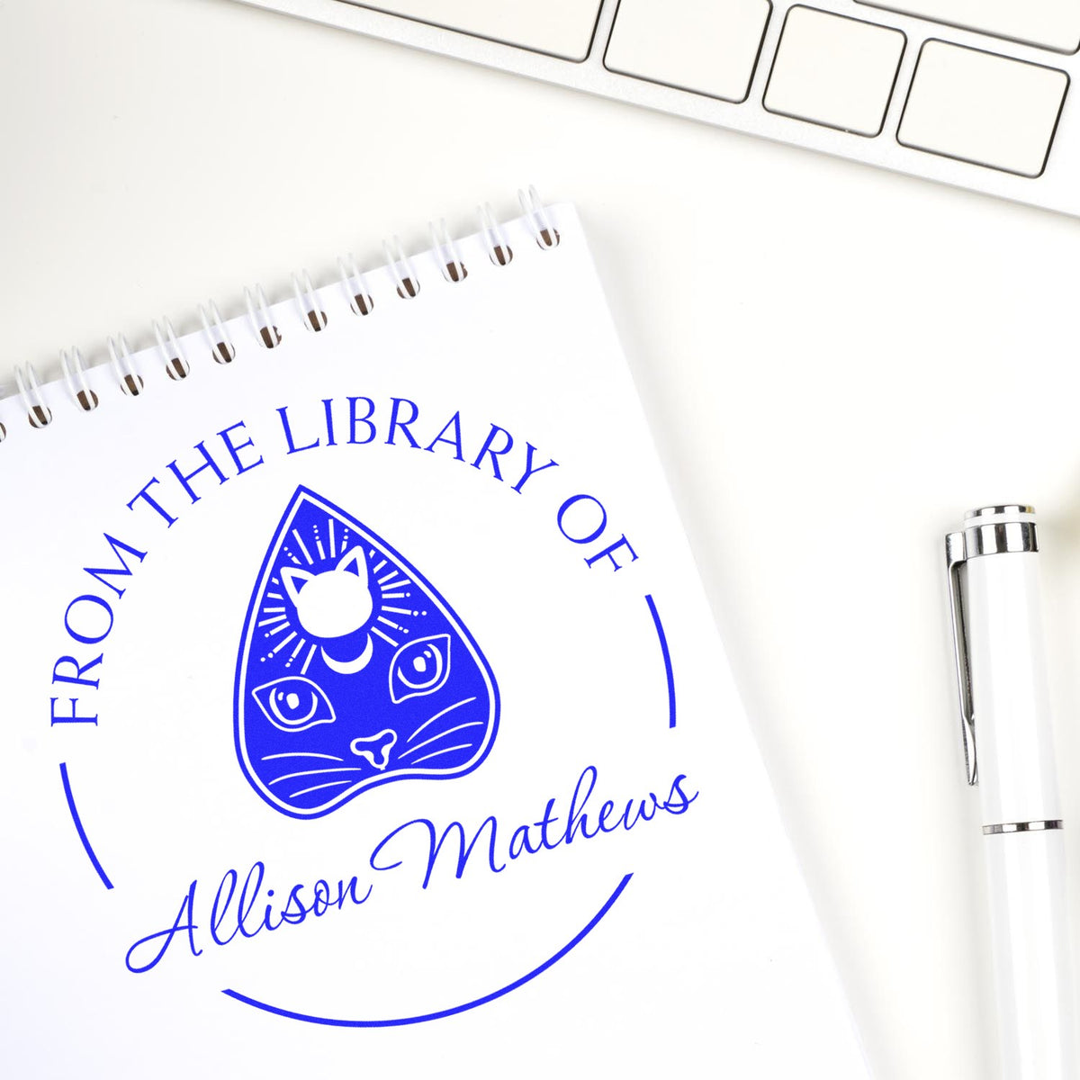 Blue custom library stamp and seal with cat logo and Allison Matthews text on a white notebook, beside a silver pen.