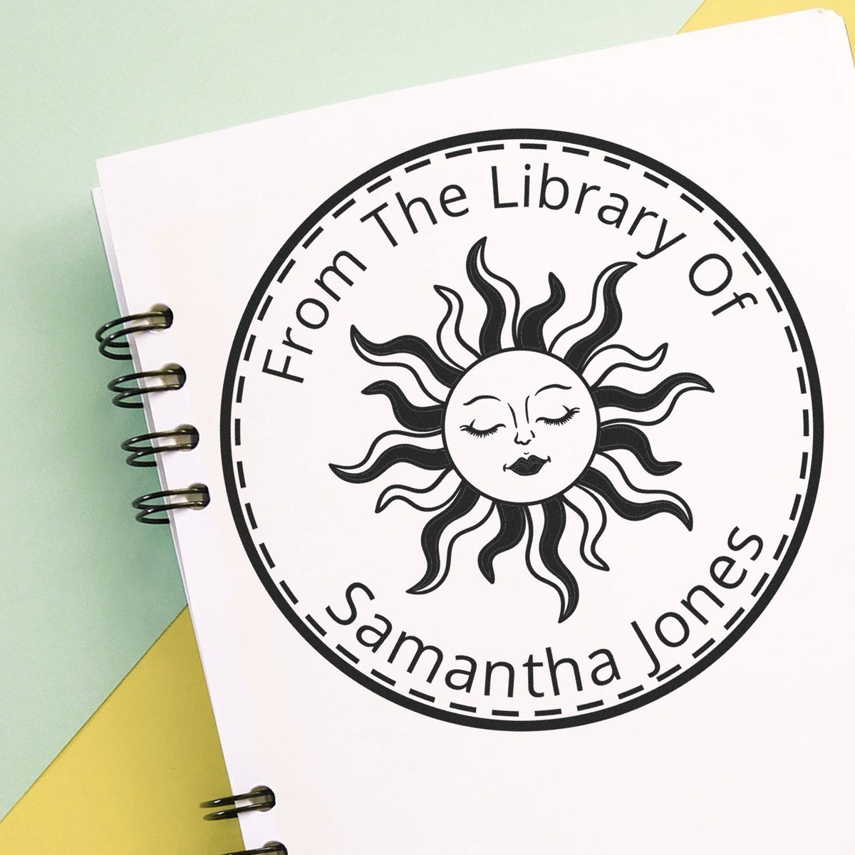 Black circular library stamp seal with sun face on white spiral notebook, reading From The Library Of Samantha Jones.