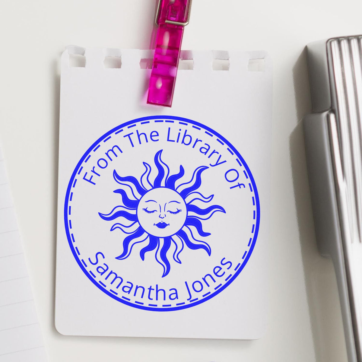Blue custom library seal stamp with sun face and pink clip on white card