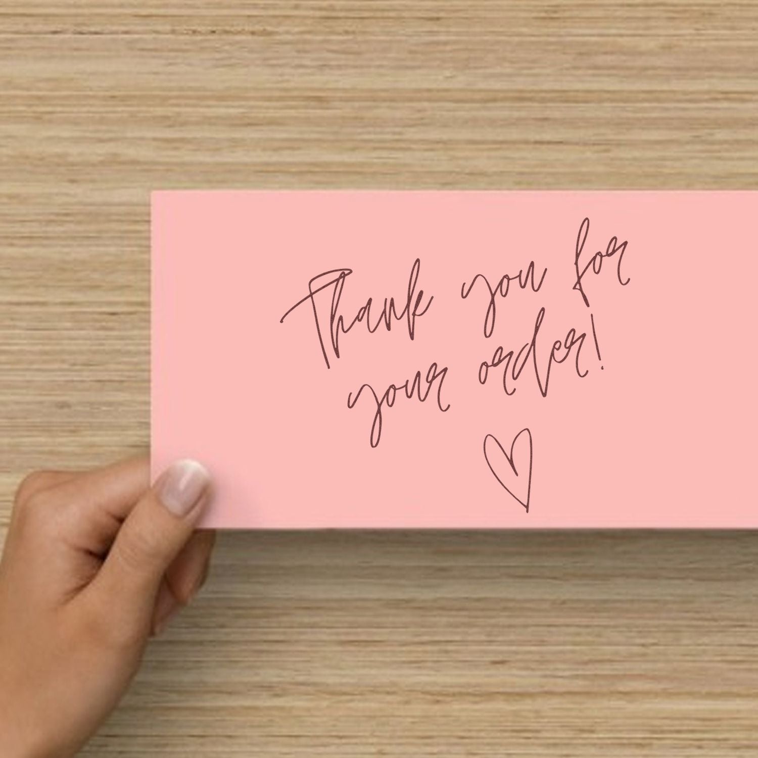 Pink thank-you stamp card with brown script and heart, held over a wood-grain table.