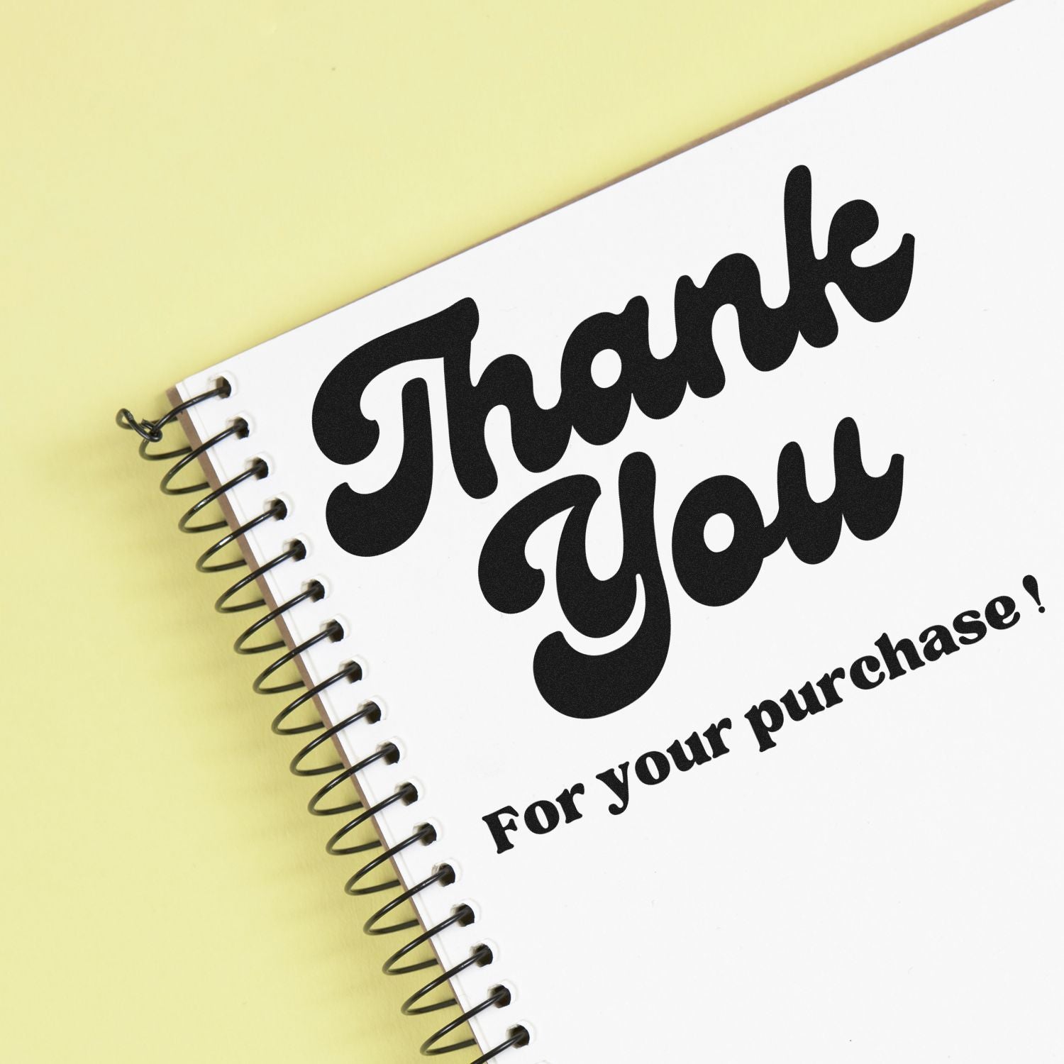 Black Thank You stamp on white spiral notebook with yellow background, reading For your purchase!