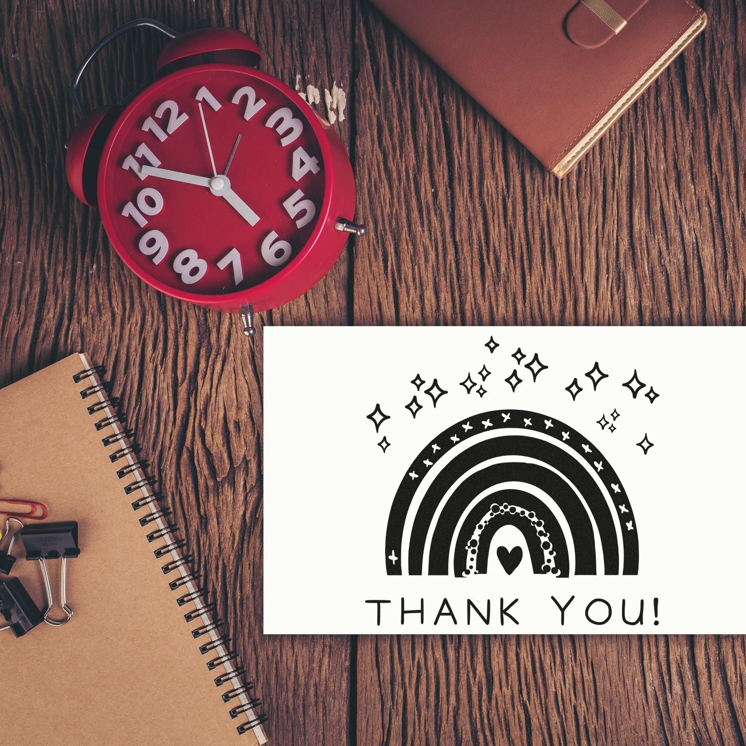 Red alarm clock, kraft notebook, and black rainbow thank-you stamp/seal on white card over wood tabletop.