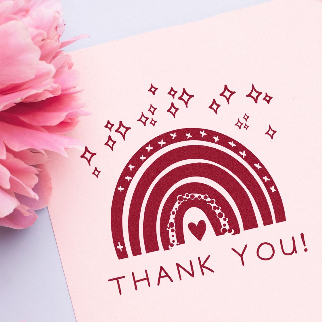 Burgundy rainbow thank-you stamp seal with heart and sparkles on pale pink paper beside pink flowers.