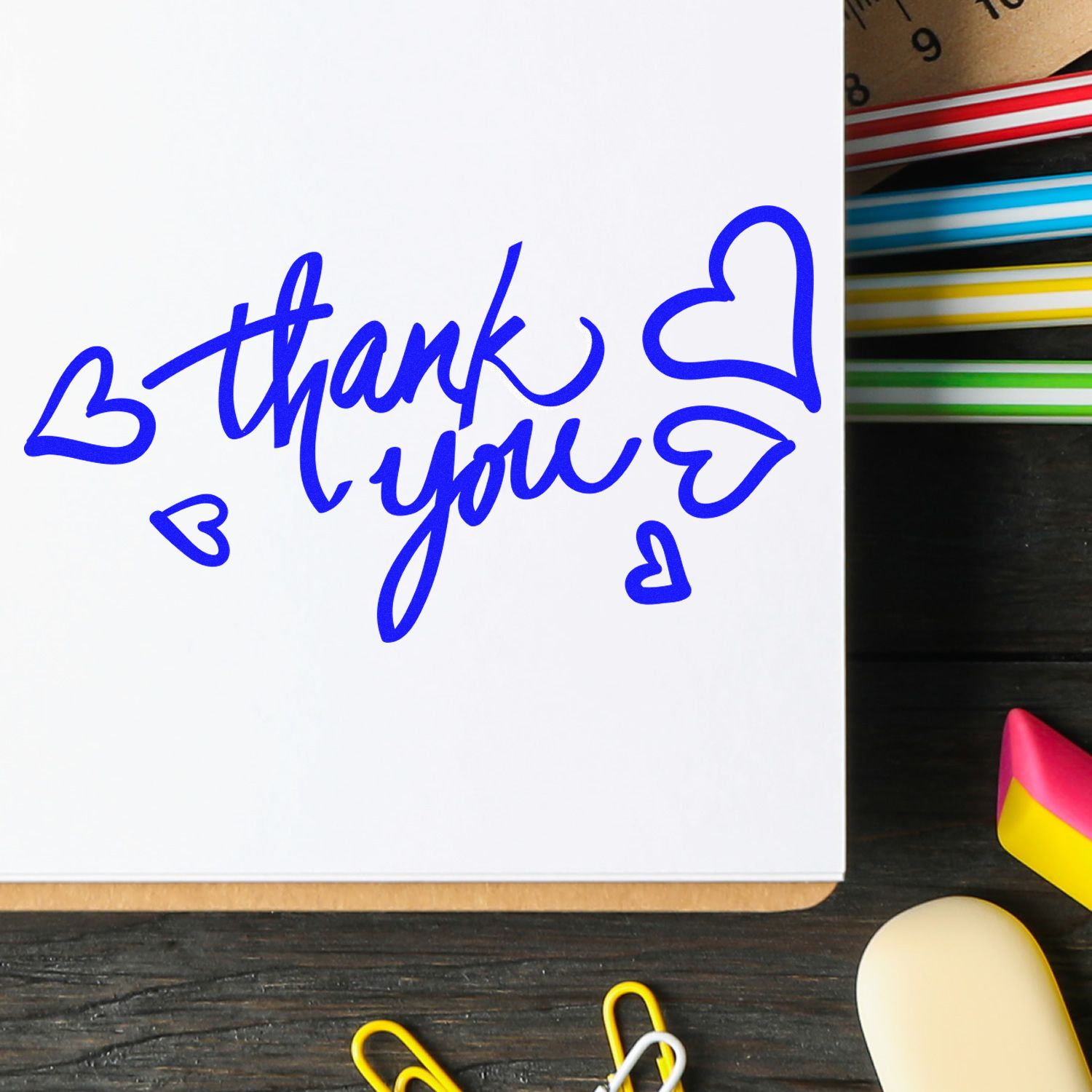 Blue handwritten thank you with hearts on white paper, beside colorful stamp and seal supplies on a dark desk.
