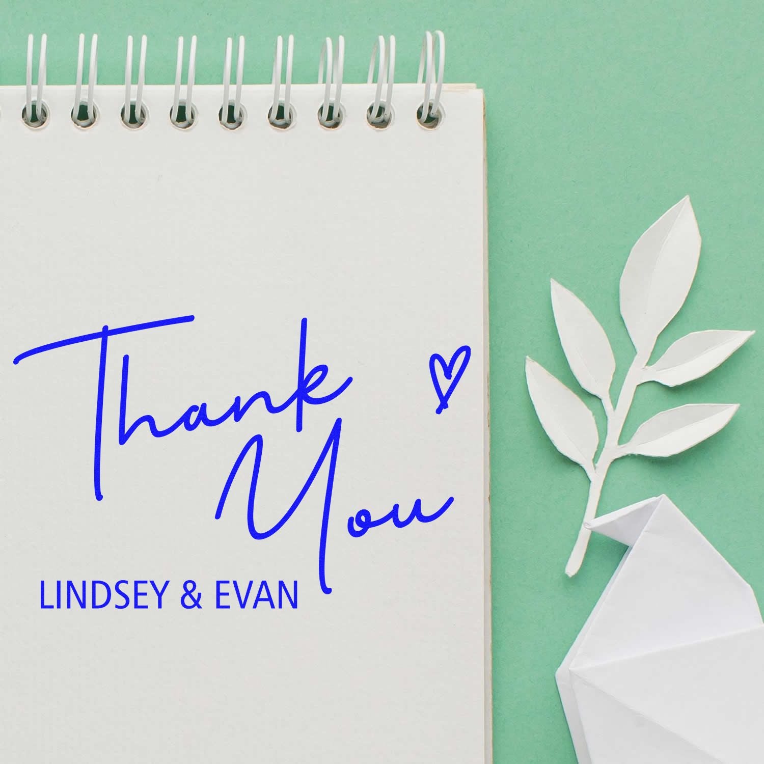 Custom blue thank-you stamp and seal set with Lindsey & Evan on a white spiral notepad, mint green background, white paper leaf and envelope.