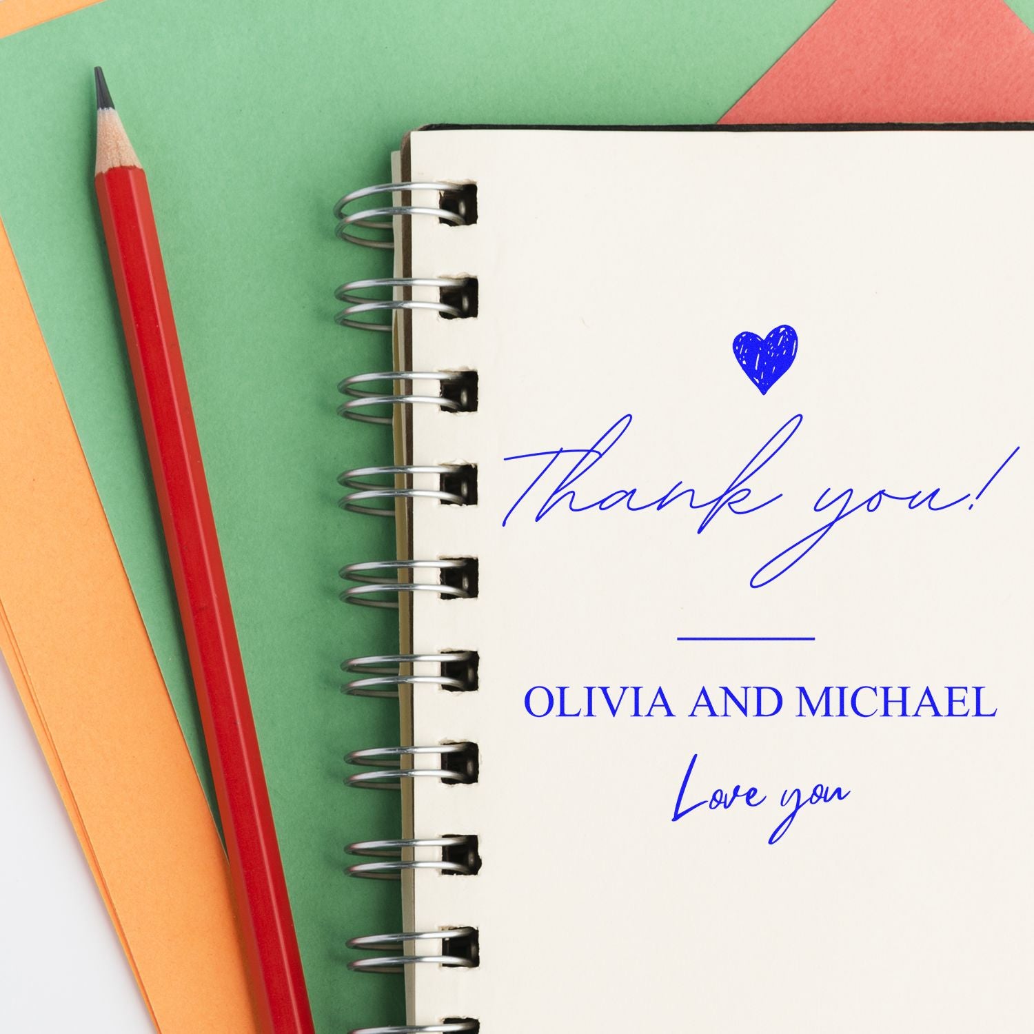 Personalized blue thank-you stamp and seal on spiral notebook, with heart, Olivia and Michael, beside a red pencil on green paper.