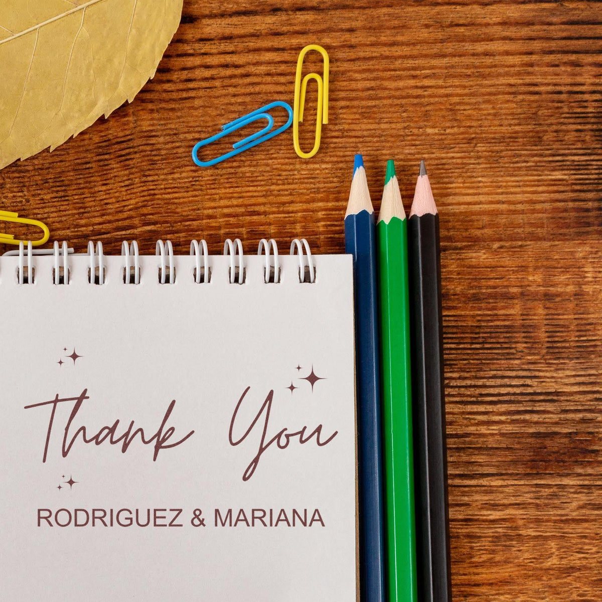 Custom thank-you stamp and seal card with Rodriguez & Mariana, brown script on white notebook, beside blue, green, and black pencils.