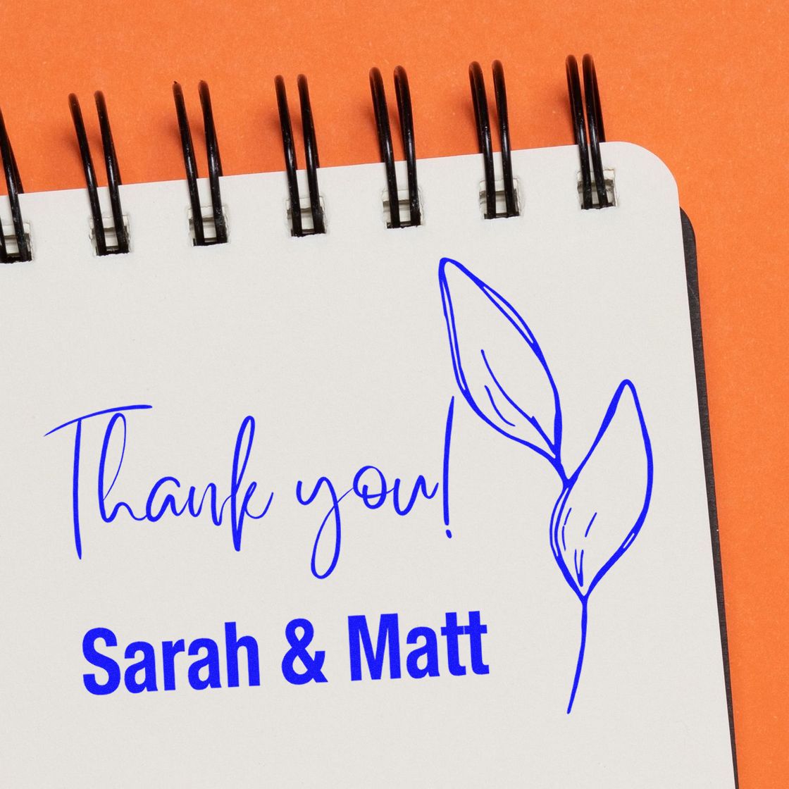 Blue custom thank-you stamp reading Sarah & Matt with leaf seal on a white spiral notebook page, set against an orange background.