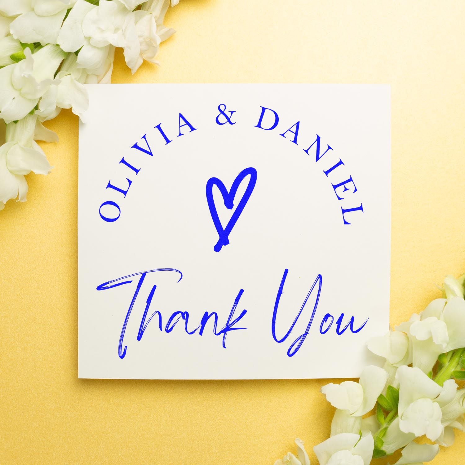 Blue custom wedding stamp and seal for Olivia & Daniel, with heart logo and Thank You on white card, pale yellow background, white flowers.
