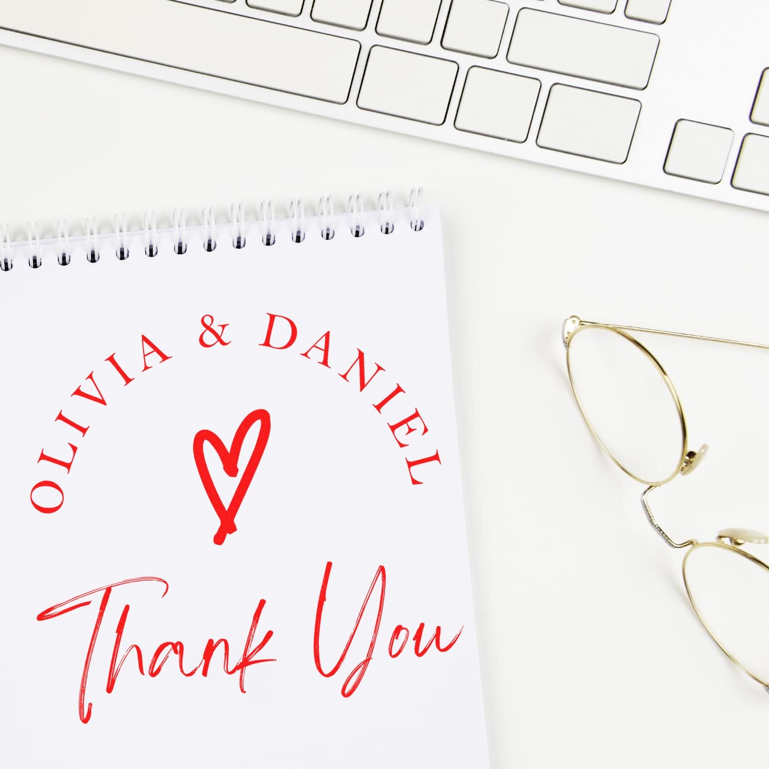 Custom red stamp and seal design on white notebook: Olivia & Daniel, heart, and Thank You beside gold glasses and keyboard