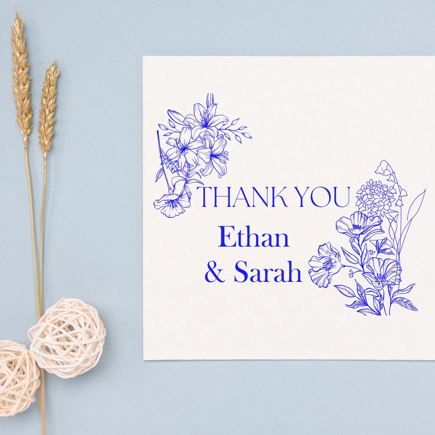 Blue floral custom stamp and seal reading Thank You Ethan & Sarah on a white card, with dried wheat and woven hearts.