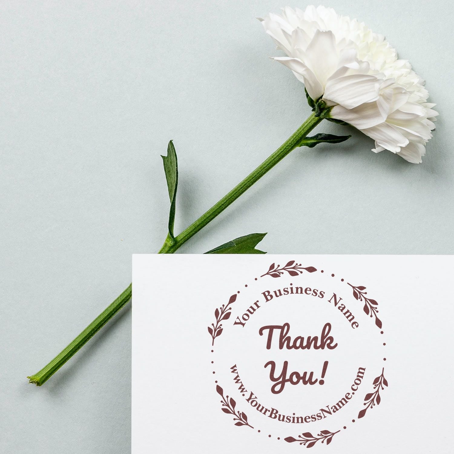 White flower beside a brown custom thank-you stamp seal on a white card, with green stem on a pale gray background.