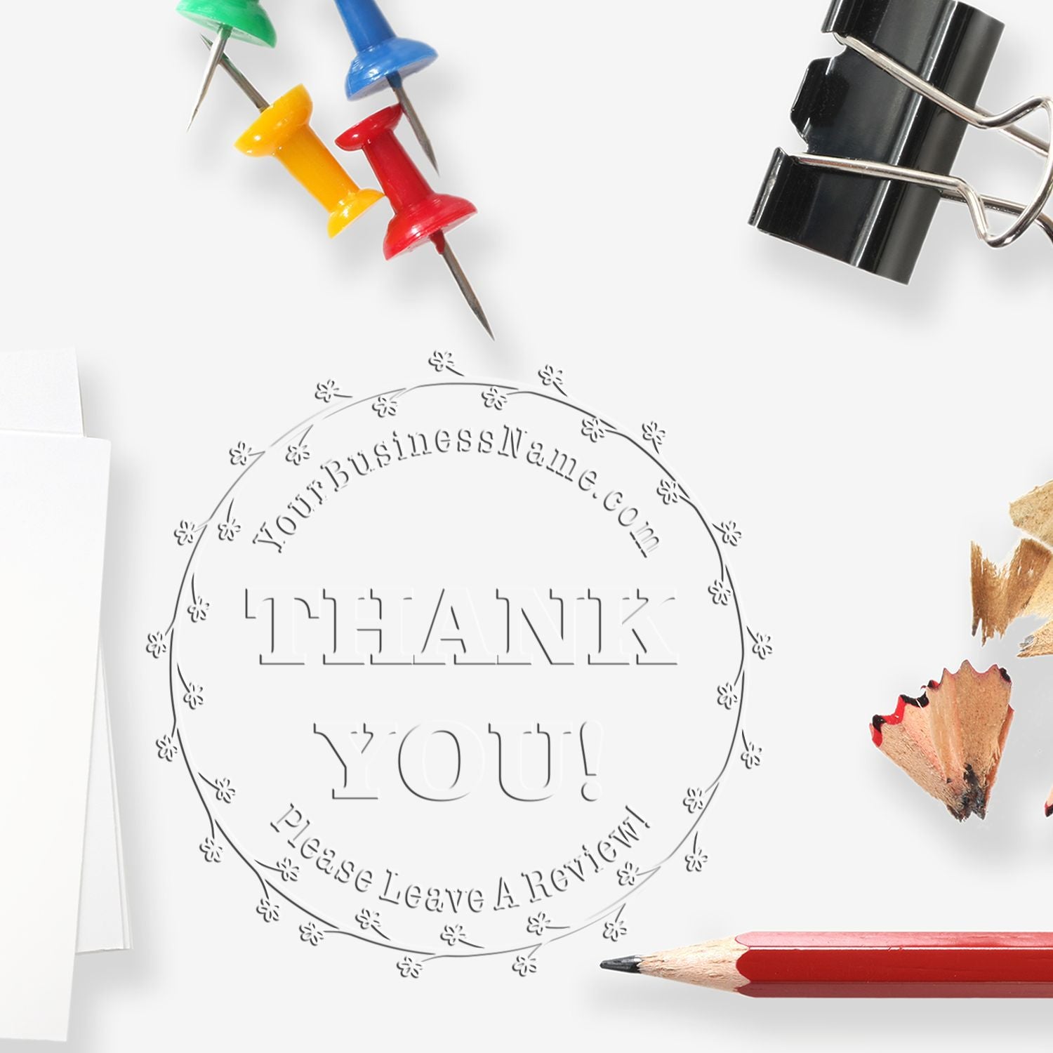 Circular thank-you embossing seal with black binder clip, colorful push pins, and white paper on a clean background