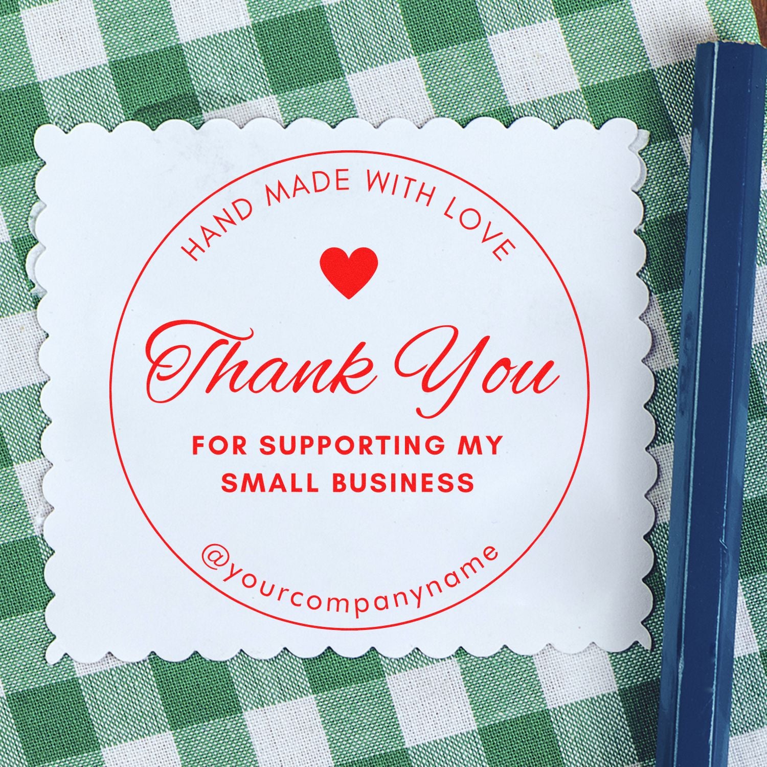White scalloped thank-you stamp seal with red heart and text on green checkered fabric, beside a navy blue packaging strip.