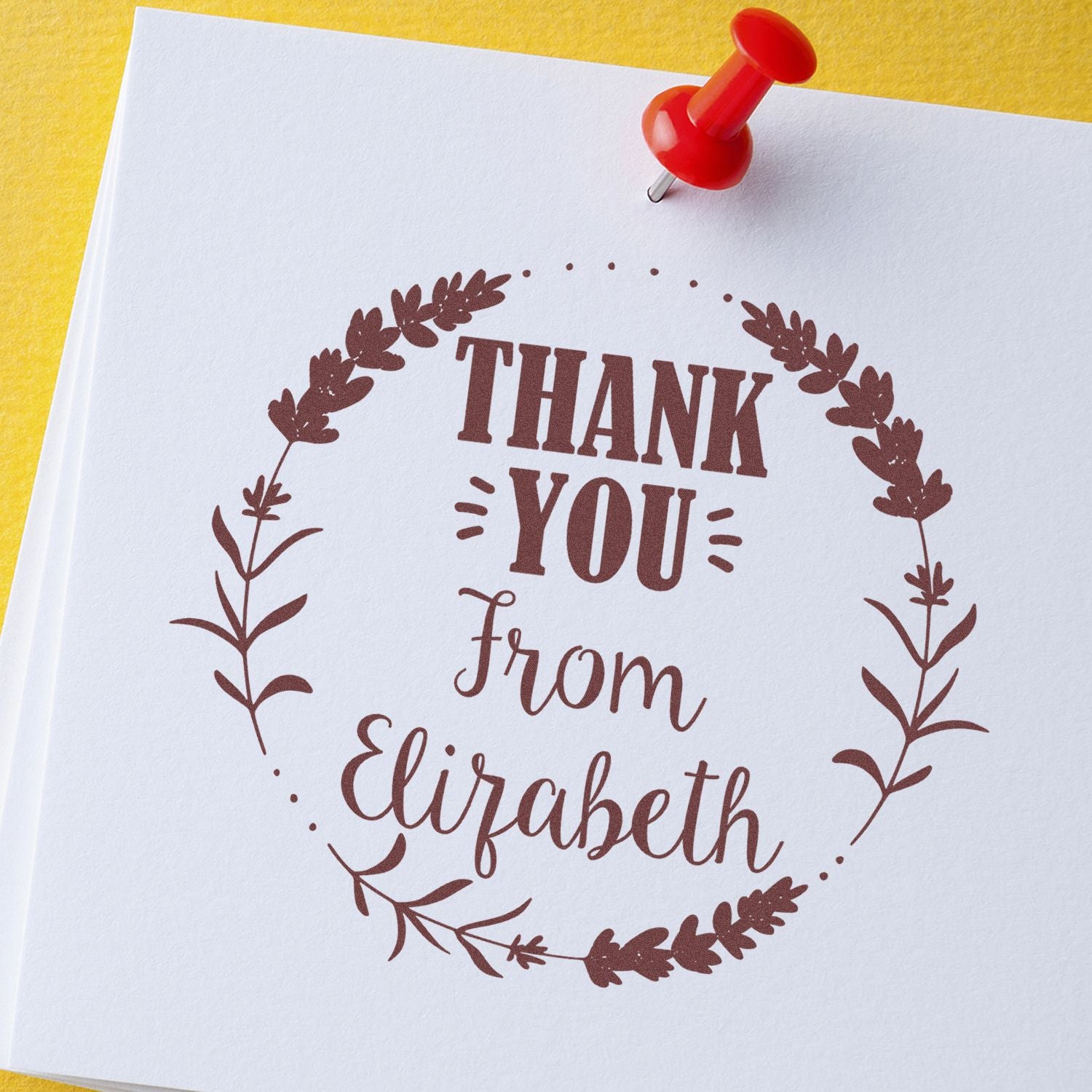 Brown custom thank-you seal stamp for Elizabeth on white paper, with floral wreath and red push pin on yellow background