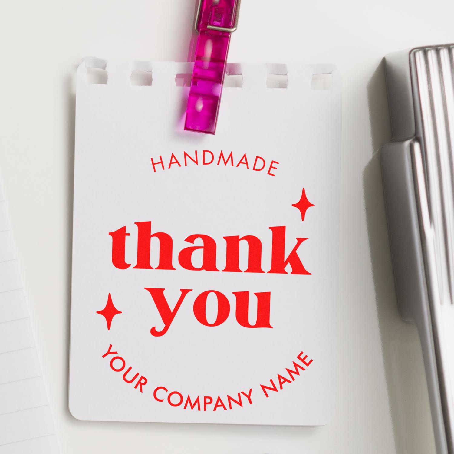 Custom red thank-you stamp seal on white card, clipped with pink clip, beside silver stamp block.