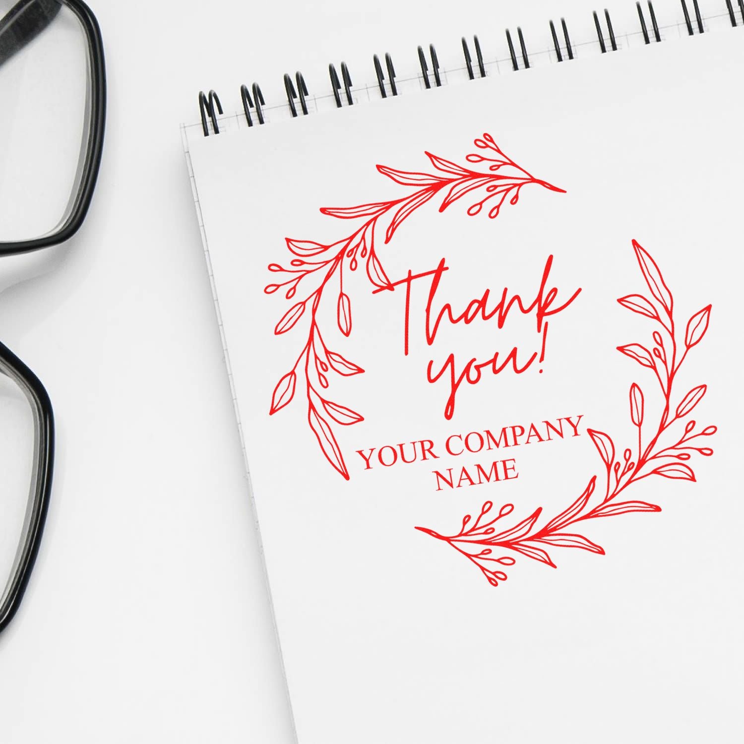 Red custom thank-you stamp seal with floral wreath and company name on a white spiral notebook, beside black eyeglasses.