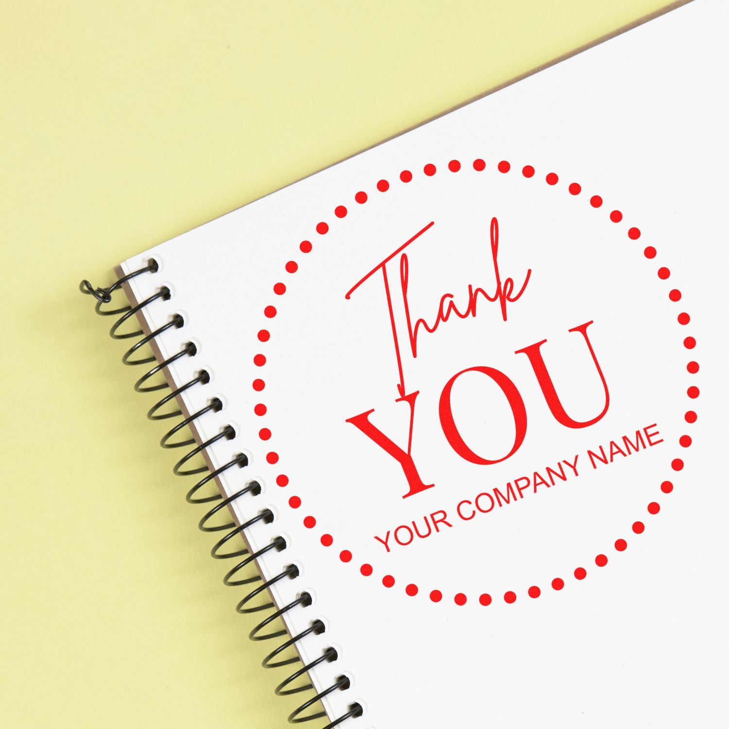 Red thank-you stamp seal with dotted circle and company name on white spiral notebook, on pale yellow background.