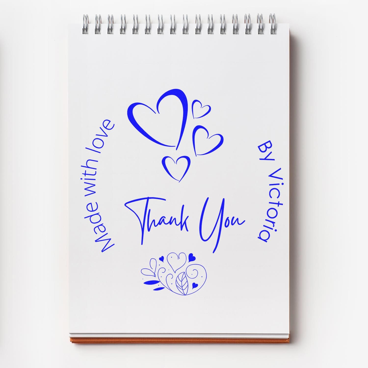 Blue stamp and seal design with hearts, Thank You, Made with love, and By Victoria on a white spiral notebook.