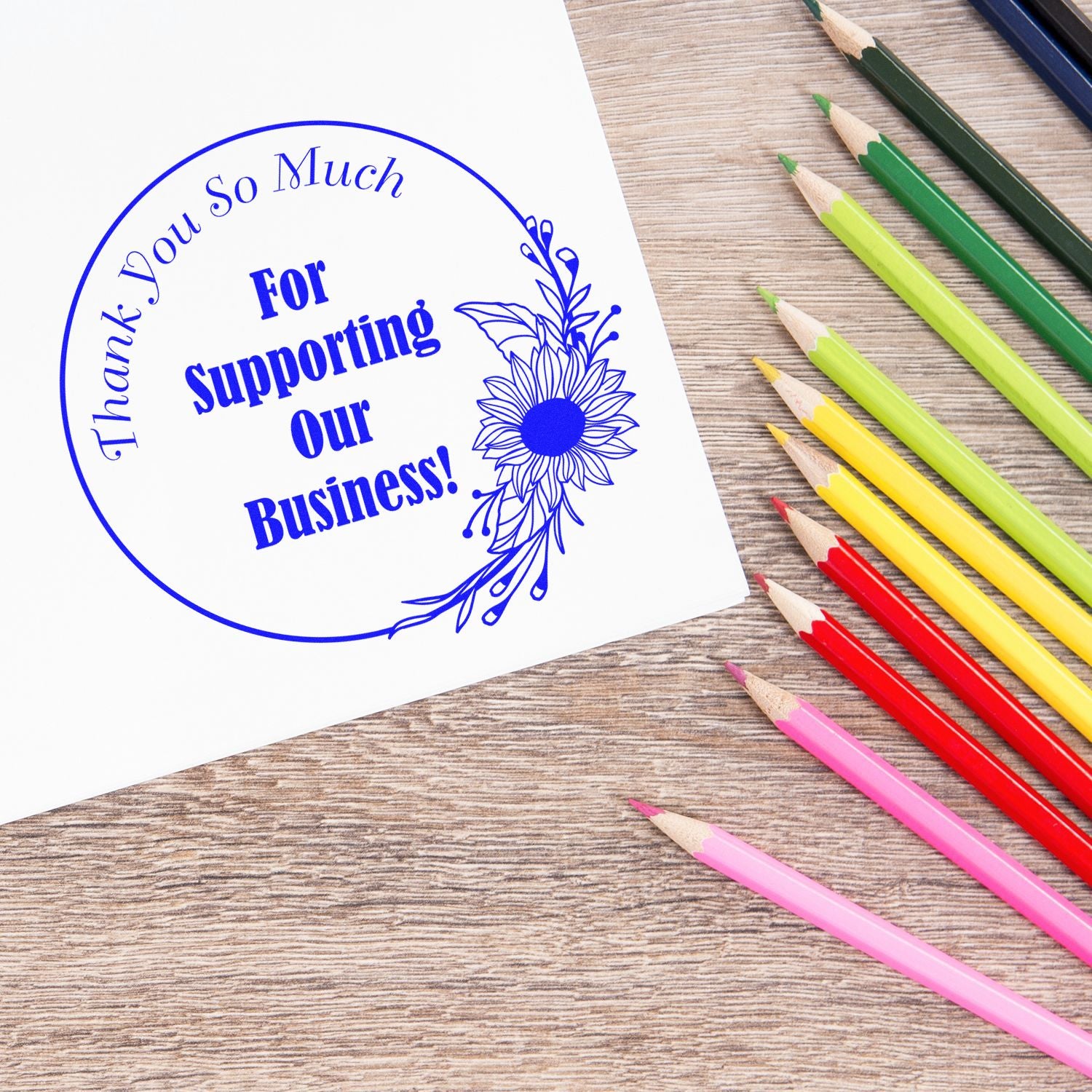 Blue custom stamp and seal saying Thank You for Supporting Our Business! beside colorful pencils on a wood desk.