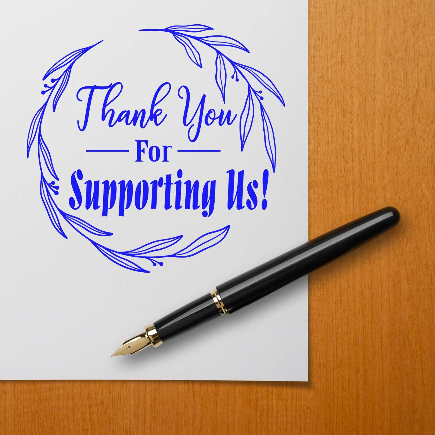 Blue thank-you stamp and seal design with leafy wreath on white paper beside a black fountain pen on a wood desk.
