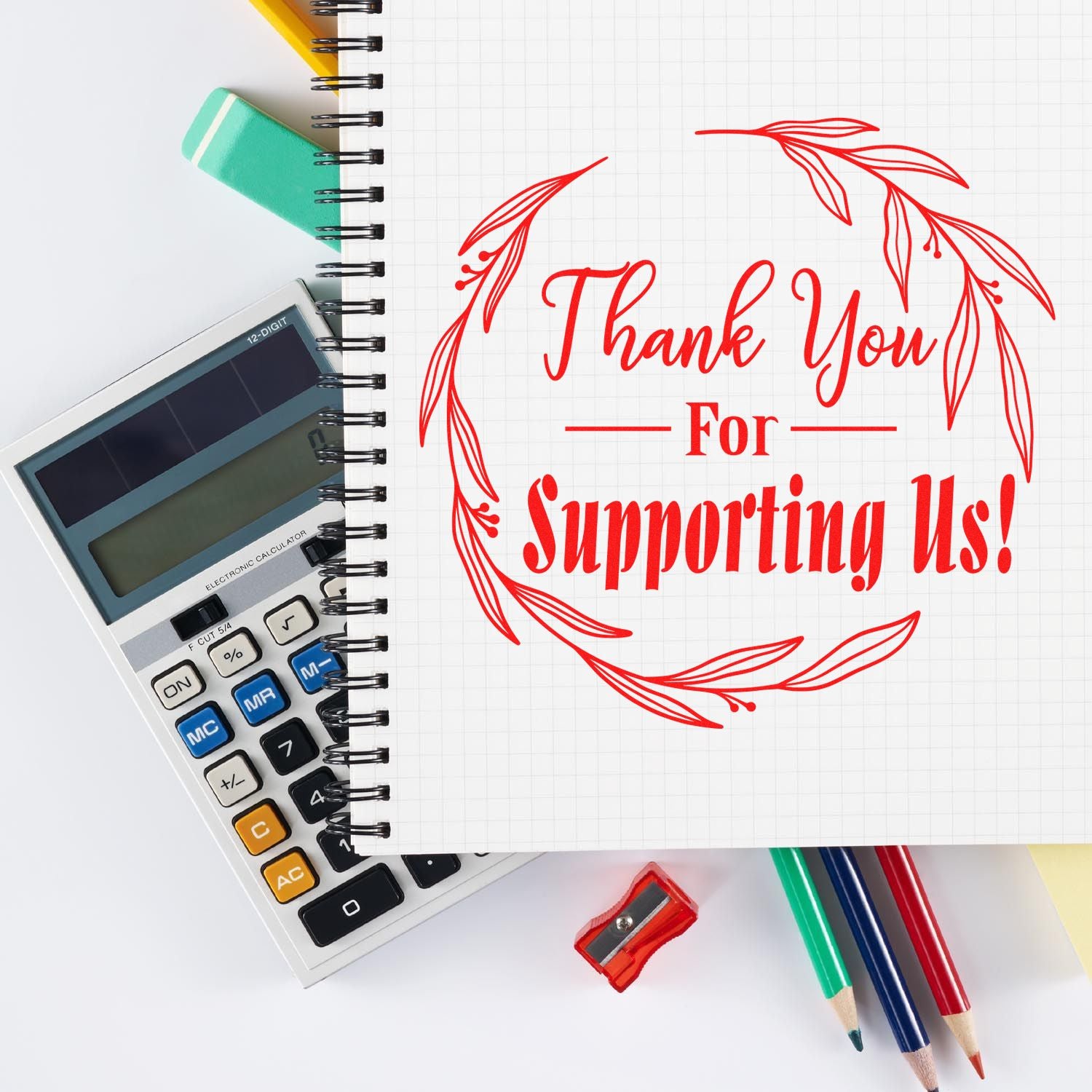 Red Thank You For Supporting Us stamp seal on spiral notebook, with calculator, eraser, pencils and sharpener on a white desk.