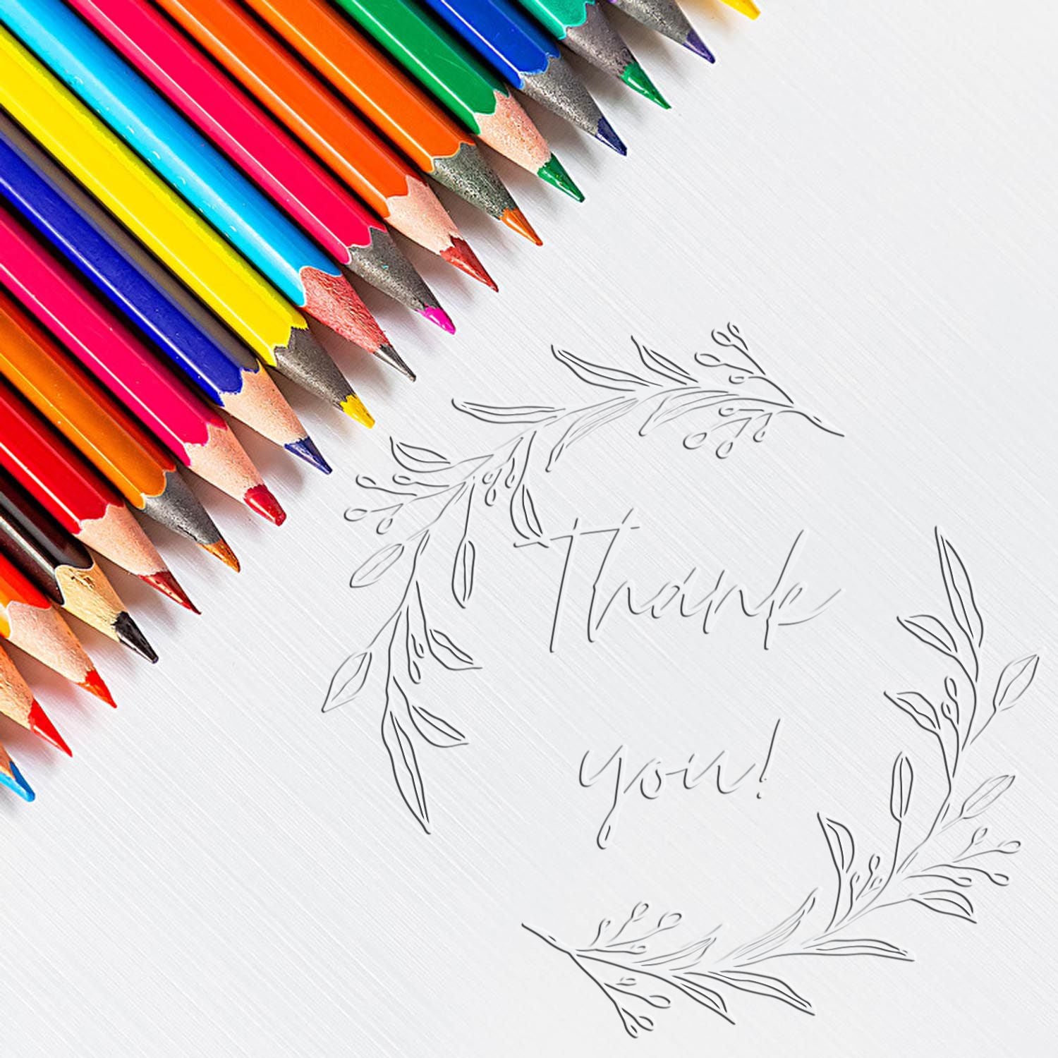 Colorful pencils beside an embossed thank you seal stamp with leafy wreath on white textured paper