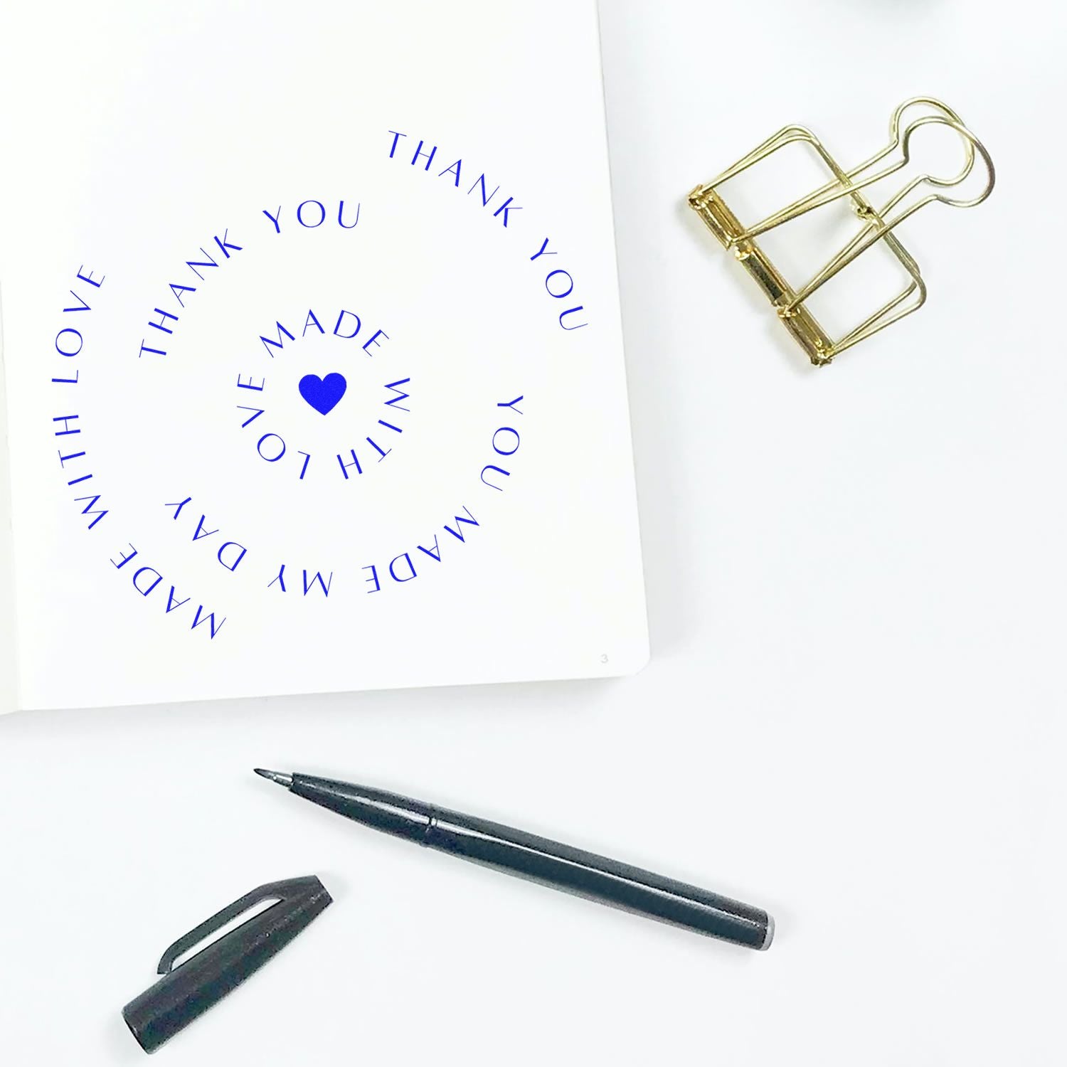 Blue spiral thank-you stamp on white card with gold binder clip, black pen and cap, showcasing custom seal products.