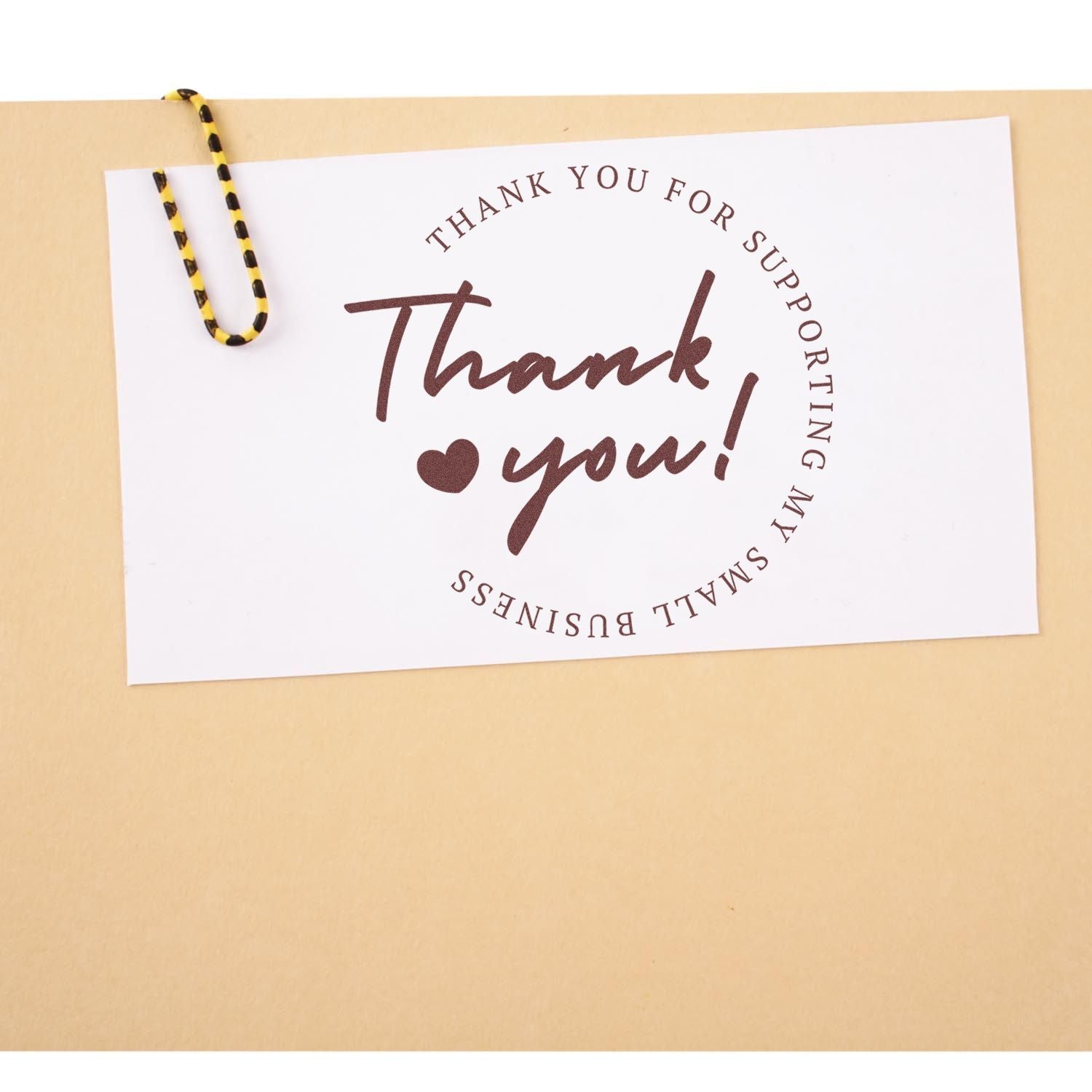 Thank-you stamp and seal on white card, brown lettering with heart, clipped by yellow-and-black paperclip on beige background.