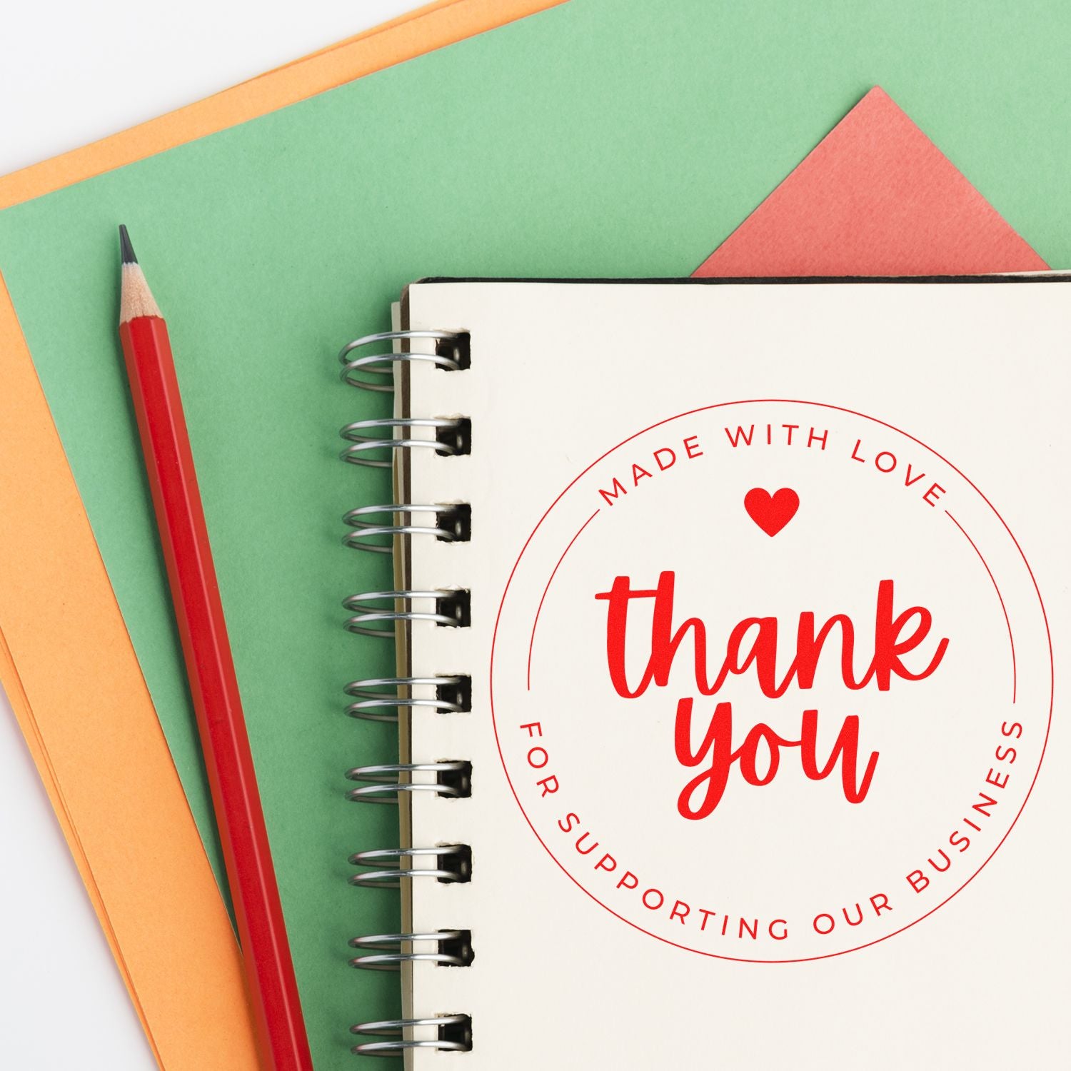 Red thank-you stamp seal on a spiral notebook beside a red pencil, set on green and orange paper backgrounds.