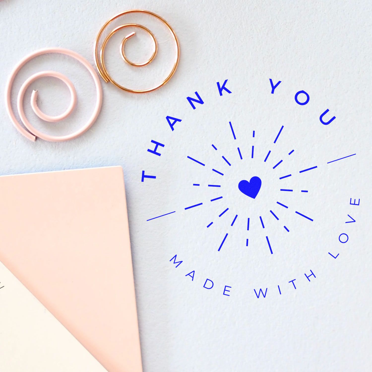 Custom stamp and seal products featuring a blue thank-you heart seal, with pink and gold spiral accents on white paper.