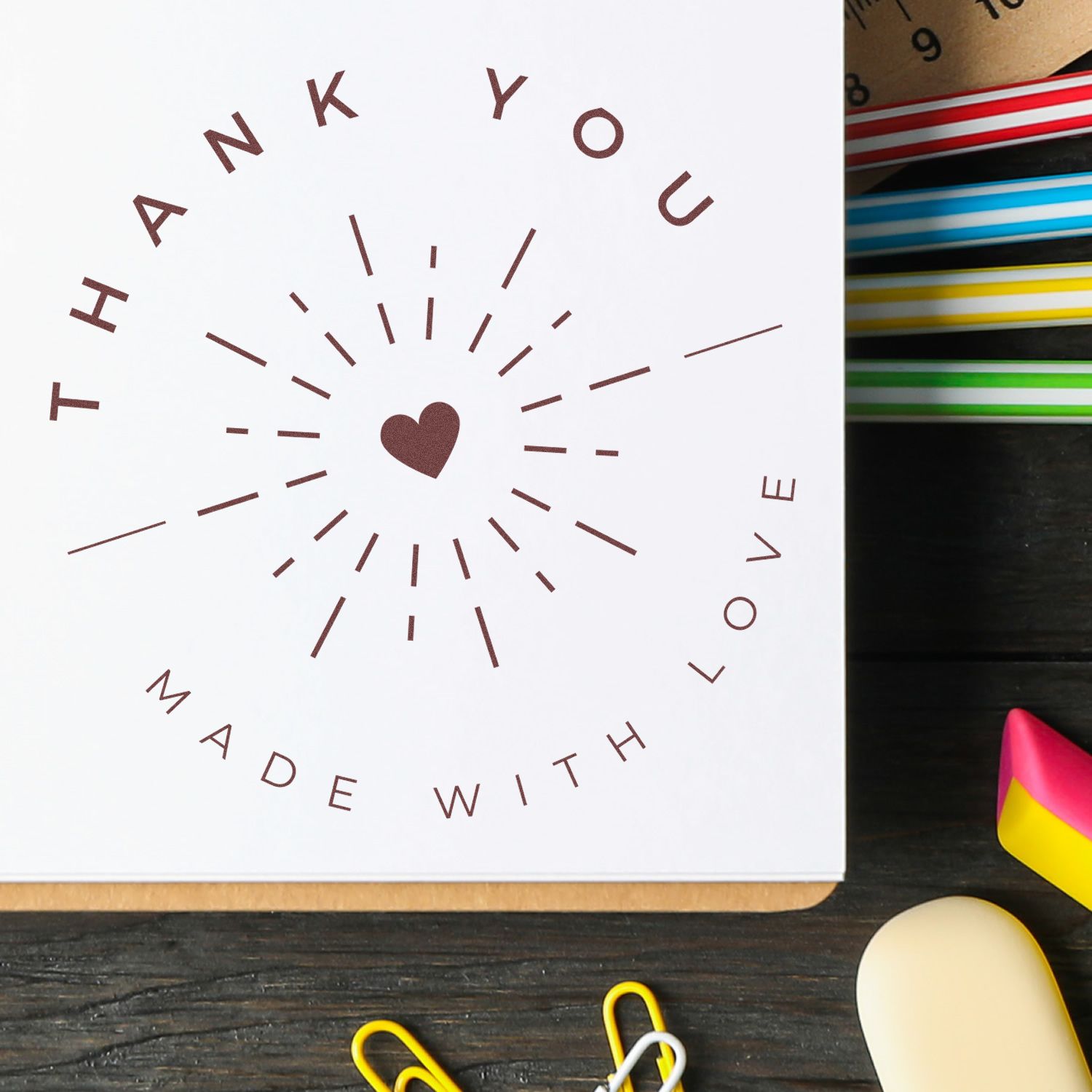 White thank-you stamp seal with brown heart and rays, surrounded by colorful ink pads on a dark desk.